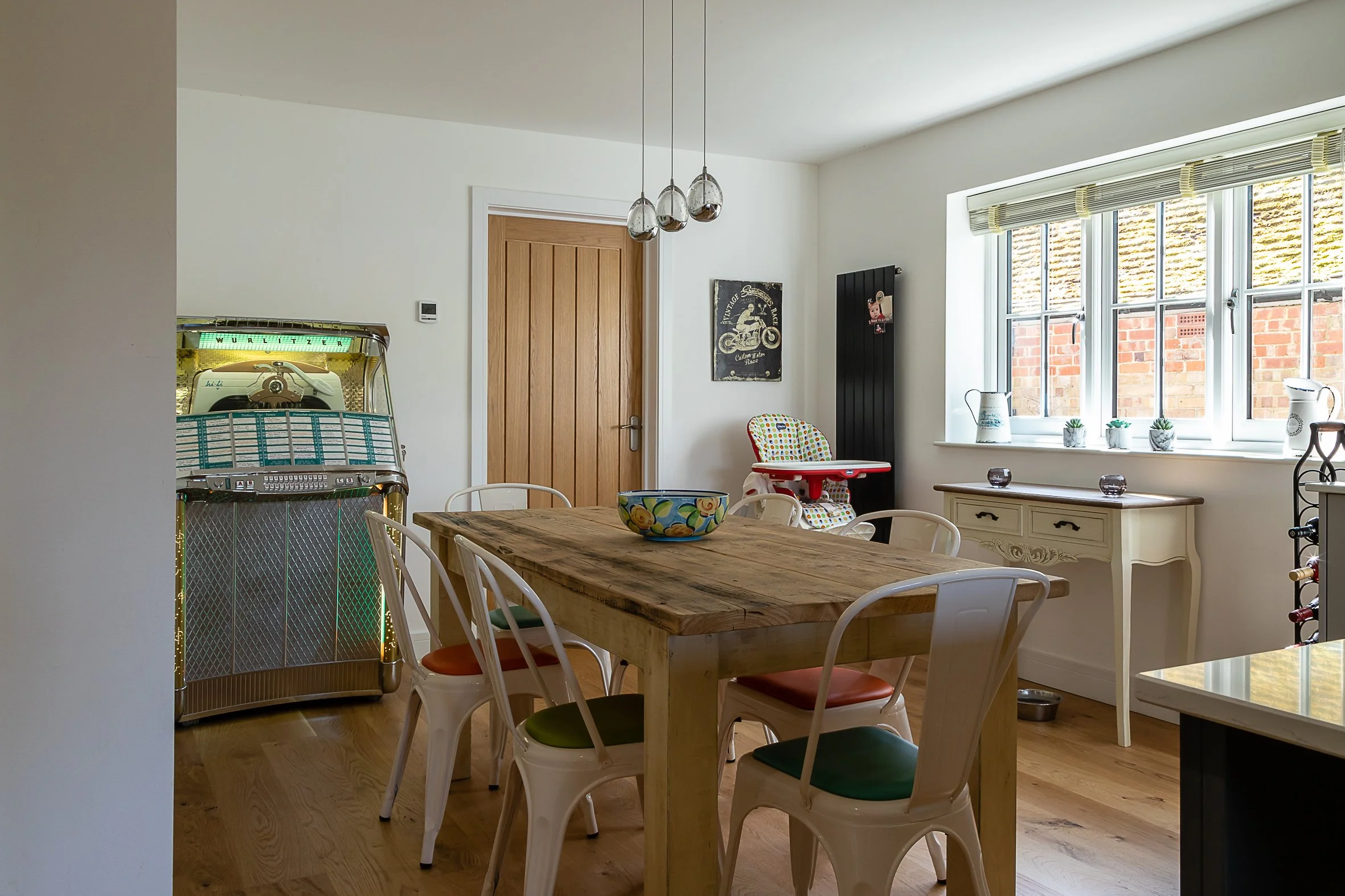 A dining area with a wooden table surrounded by six white chairs, a vintage jukebox in the corner, a high chair near a window, and a small white side table with decorative items, bright natural light coming through large windows.
