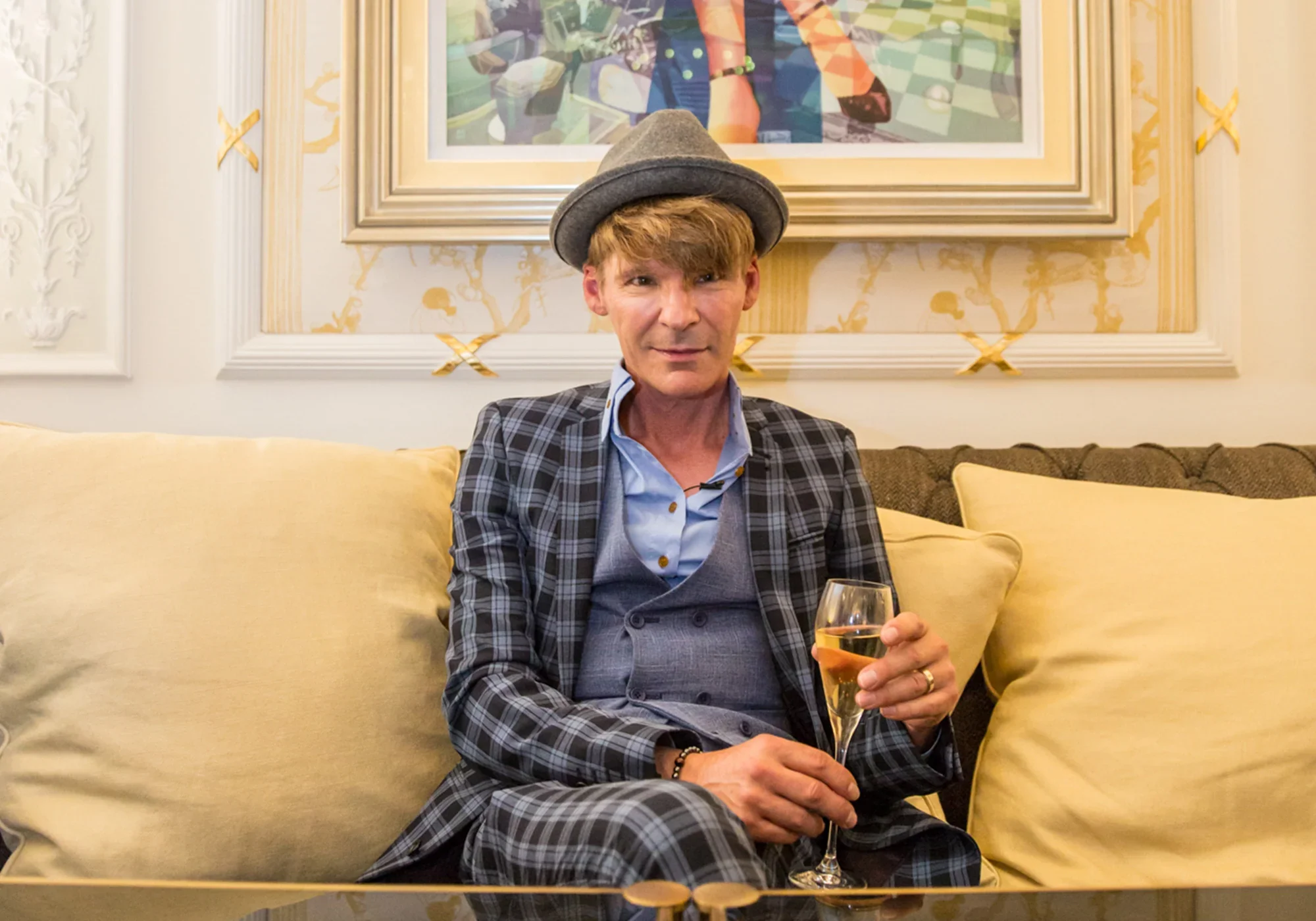 A man with reddish hair wearing a hat and a suit sitting on a yellow couch, holding a glass of champagne. Behind him is a framed colorful abstract painting and ornate wall decor.