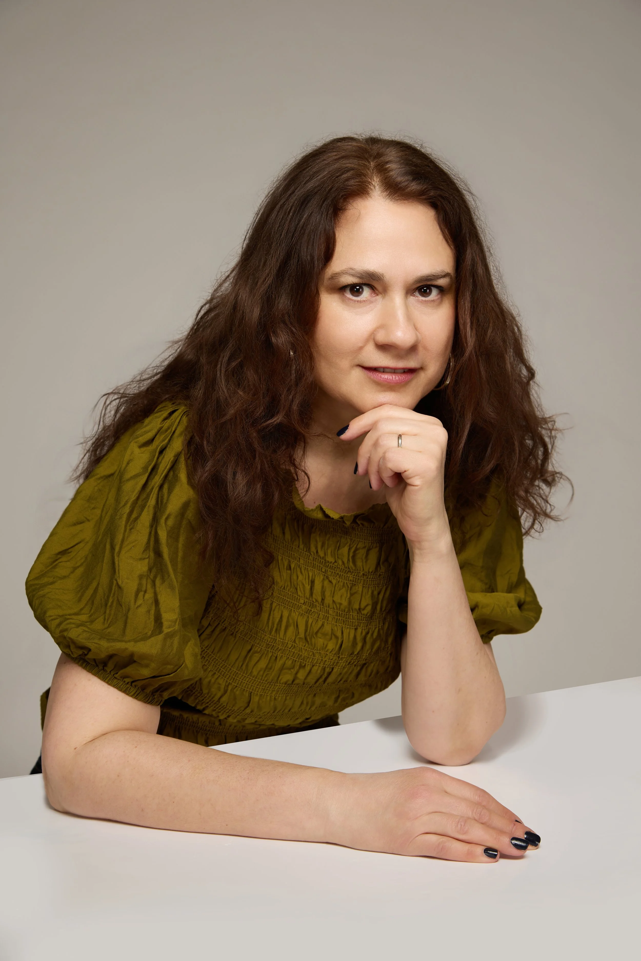 A woman with brown, curly hair and fair skin, resting her chin on her hand, wearing a green puff-sleeve blouse, seated at a white table against a neutral background.