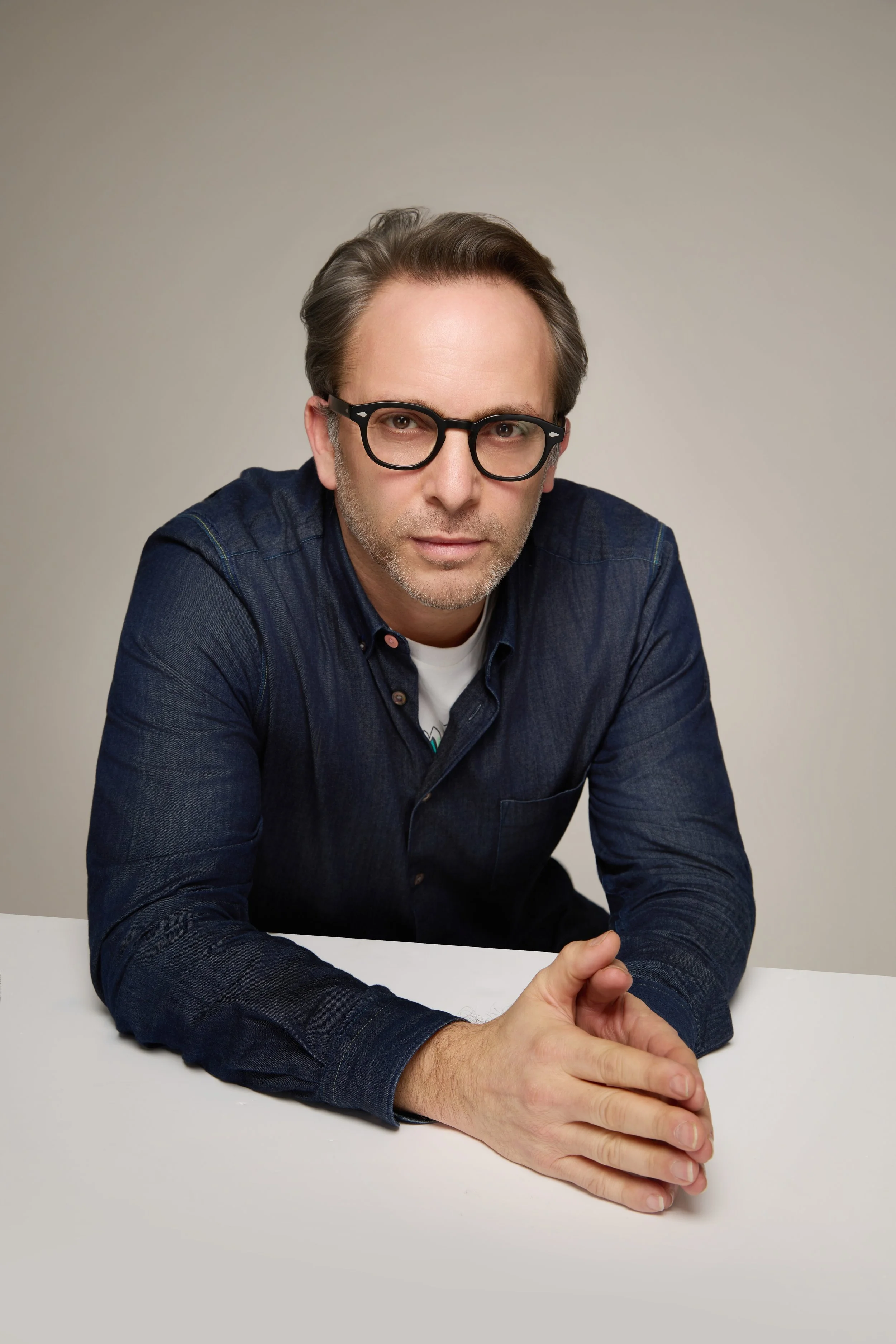 A man with gray hair, glasses, wearing a navy blue shirt, sitting at a white table with his hands clasped.