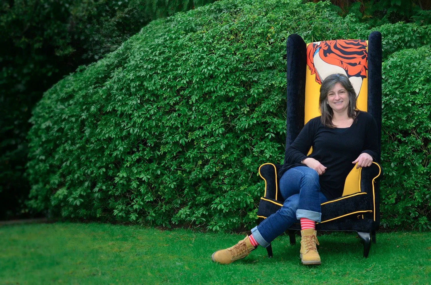 Woman sitting on a black and yellow velvet throne-like chair outdoors, with lush green bushes in the background, wearing a black top, rolled-up jeans, striped socks, and tan boots, smiling.