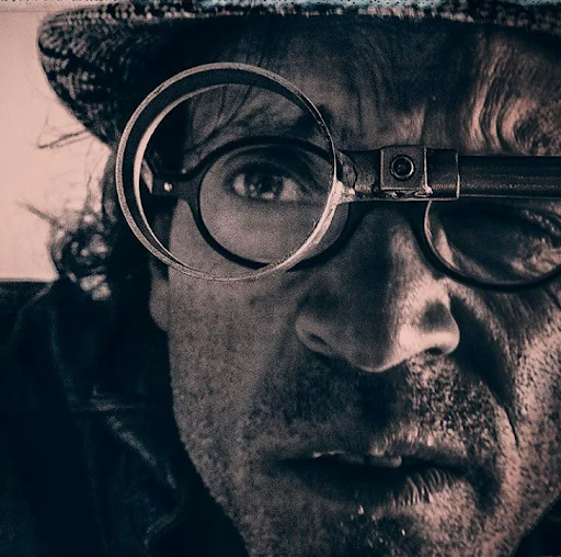 Close-up of a man wearing a hat and glasses, with one eye looking through a magnifying glass.