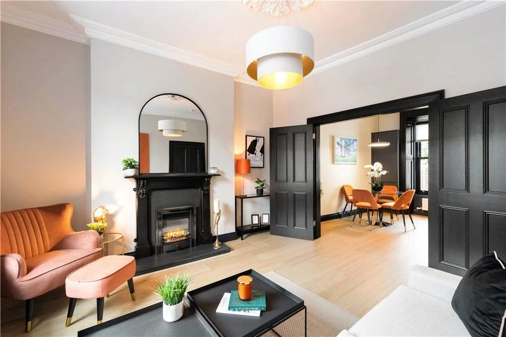 A modern living room with a fireplace, black mantel, and a large arched mirror above it. The room has a peach-colored sofa, a matching ottoman, and a glass side table with a lamp. In the background, a dining room with orange chairs around a round table and a large window is visible. The space features light hardwood flooring and white walls with crown molding.