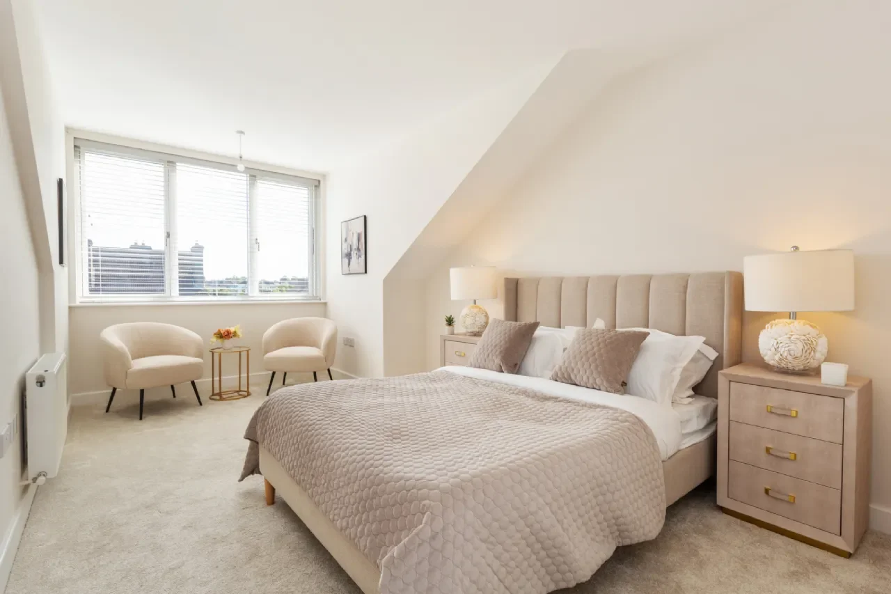 A modern bedroom with a large bed, beige headboard, nightstands with lamps, two armchairs near a window, and soft natural lighting.