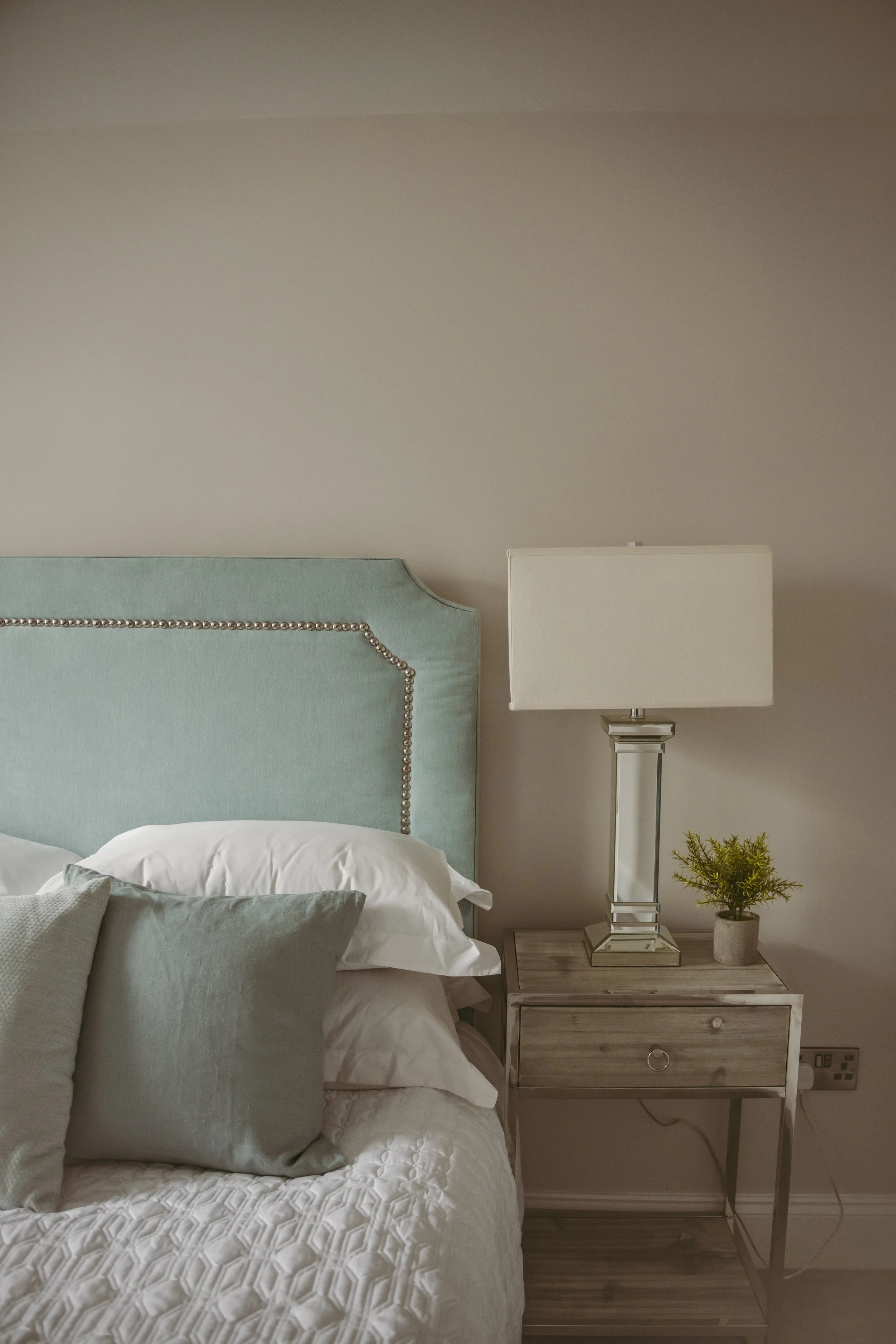 A bedroom with a bed that has a pale blue upholstered headboard with a nailhead trim. There are white and gray pillows on the bed. Next to the bed is a small bedside table with a glass and metal lamp and a small potted plant. The wall behind the bed is painted a light neutral color.