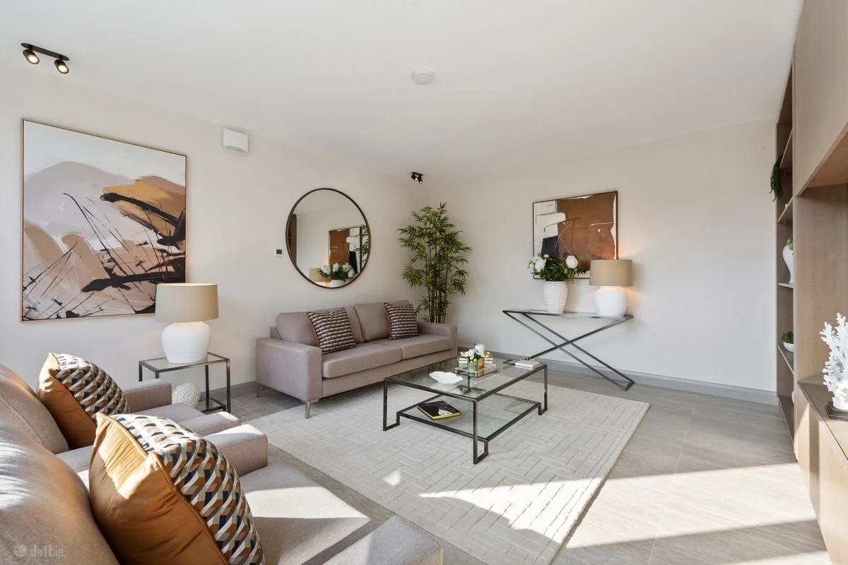 Modern living room with two beige sofas, decorative pillows, a glass coffee table, and artwork on the walls.