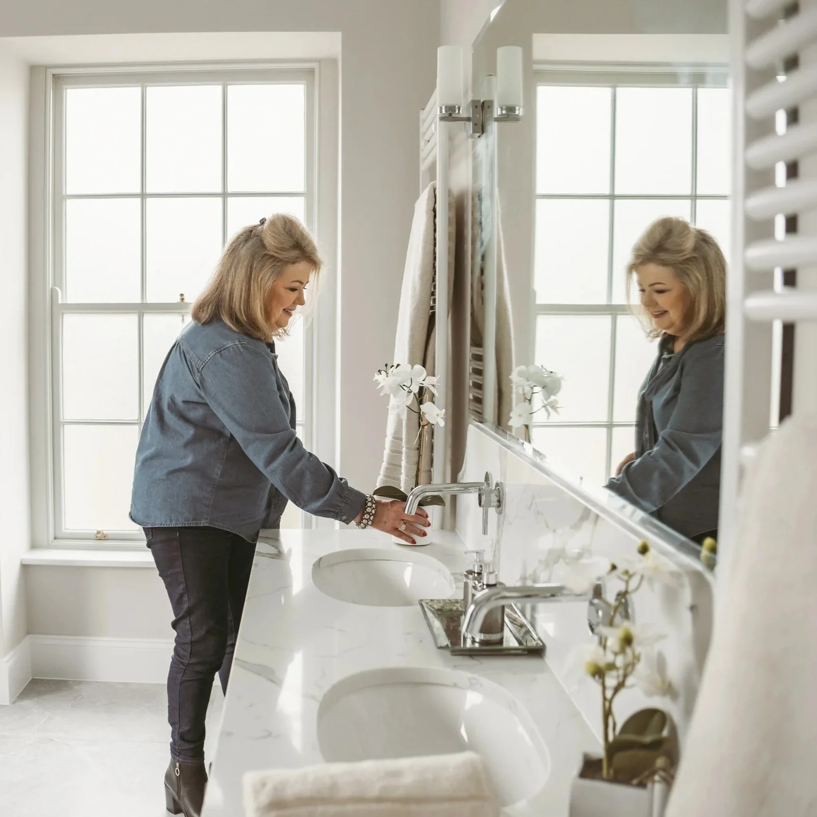A woman with shoulder-length blond hair in a denim jacket washing her hands in a bathroom sink, with a large mirror, white countertops, and a window in the background.