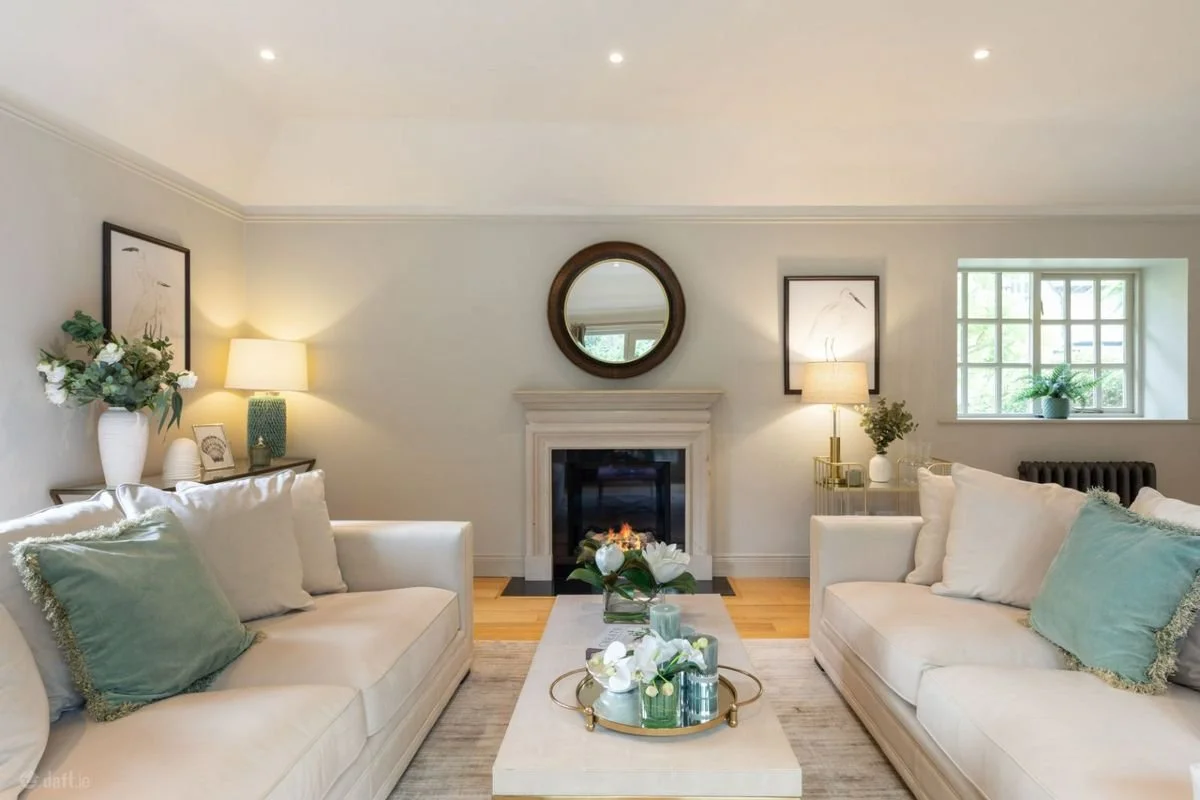 Living room with two white sofas, a fireplace with a round mirror above it, side tables with lamps, framed artwork, and a window with a plant