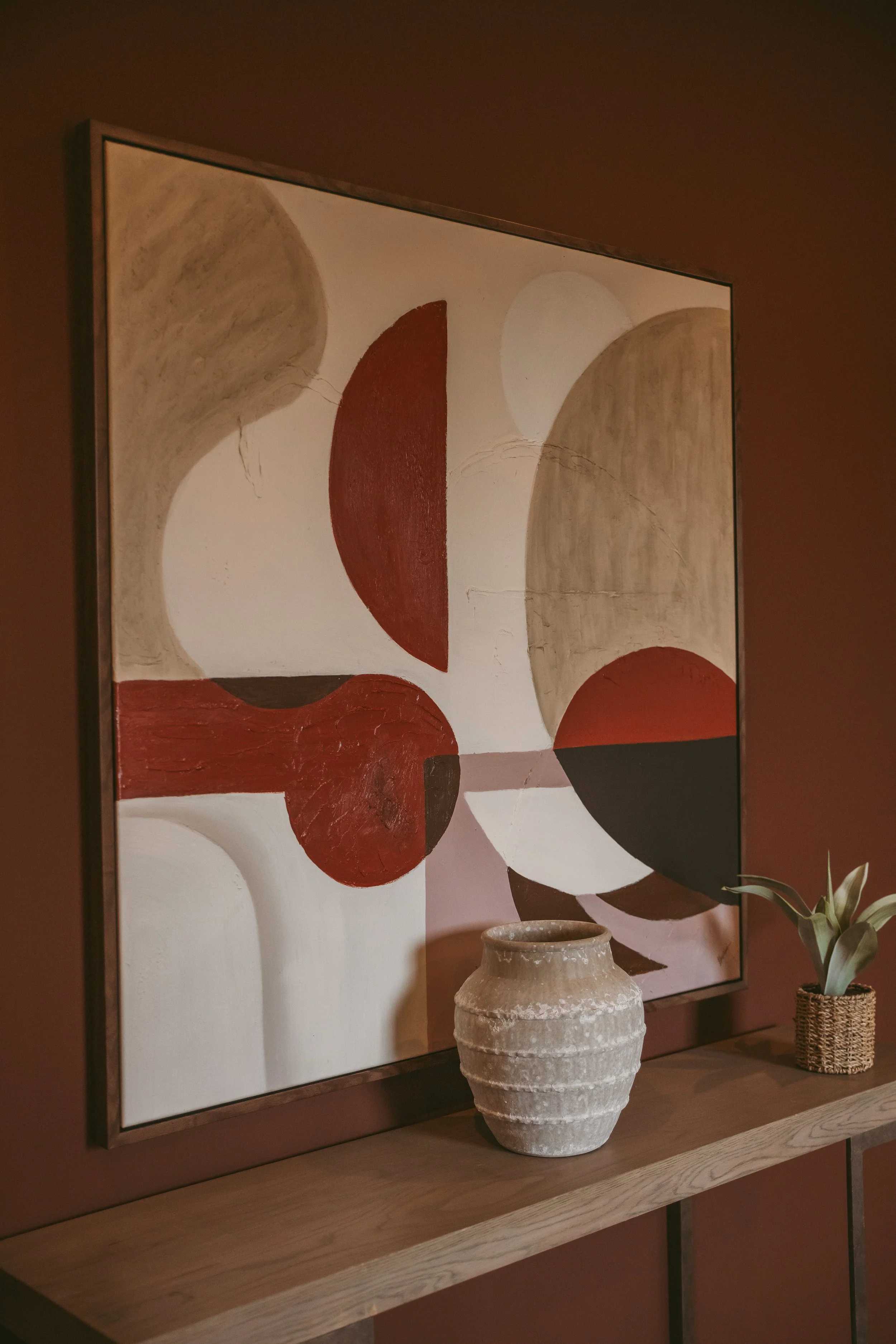 Interior decor featuring an abstract painting with geometric shapes, a large textured ceramic vase on a wooden shelf, and a potted plant.