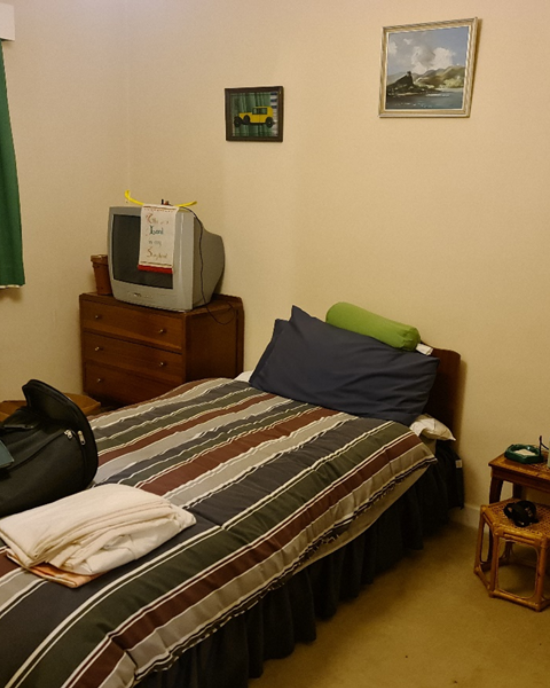A bedroom with a bed covered by a striped comforter, a dark gray pillow, and a green bolster pillow. There is a wooden nightstand with a wicker basket, and a small wooden dresser with an old-style TV on top. The TV has a note attached to it. The wall has two framed paintings, one with a landscape and the other with a yellow vehicle, possibly a jeep, with books visible in the background. A green curtain is partially visible on the left side of the image.