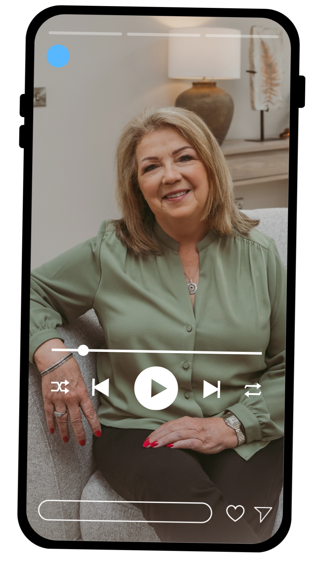 A woman with shoulder-length blonde hair smiling and sitting on a light-colored sofa in a modern, softly-lit room, wearing a light green blouse, black pants, bracelets, a watch, and a necklace, with a lamp and decorative art in the background.