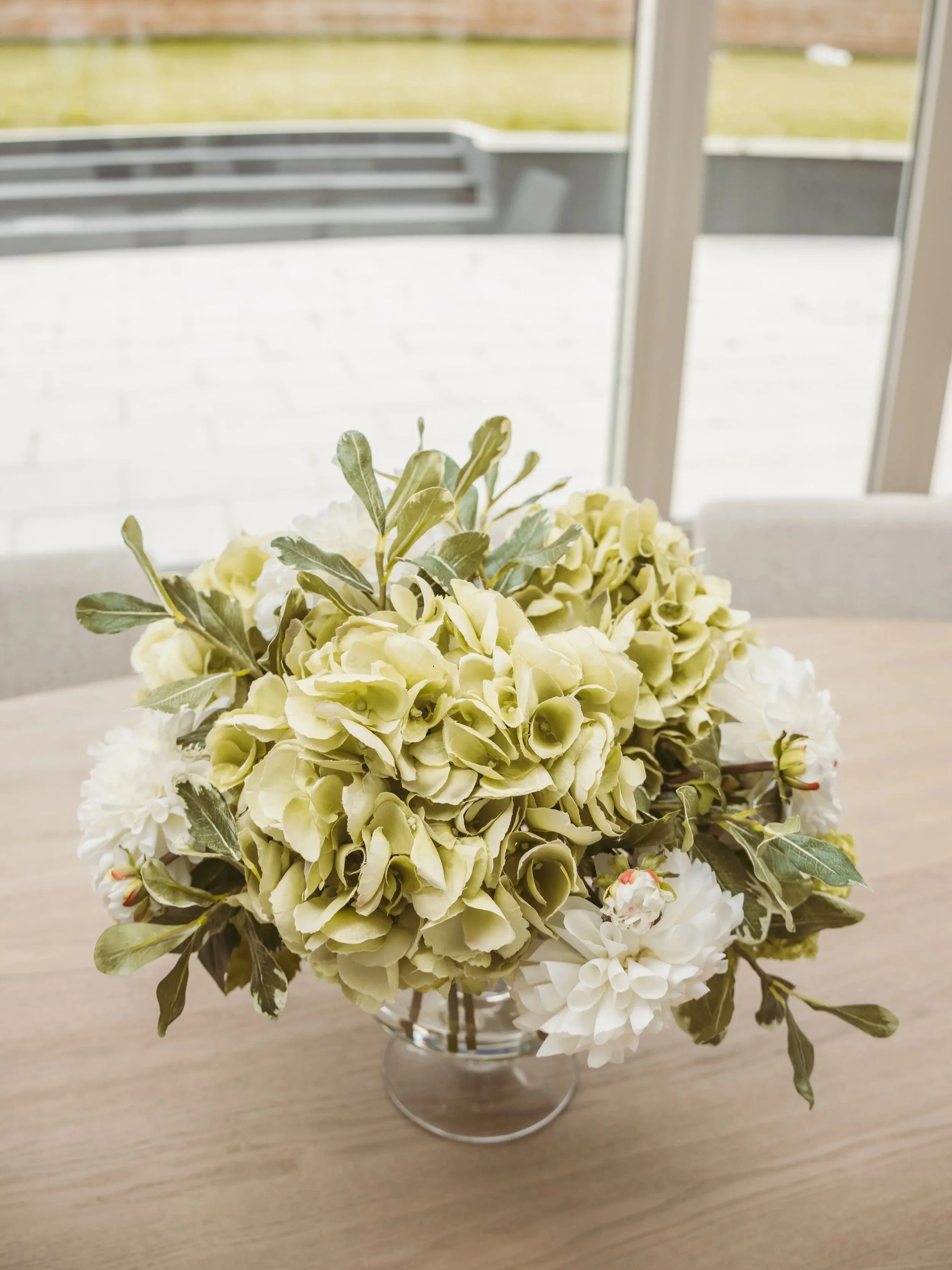 A bouquet of cream and white flowers with green leaves in a glass vase on a table, with a window and outdoor area in the background.