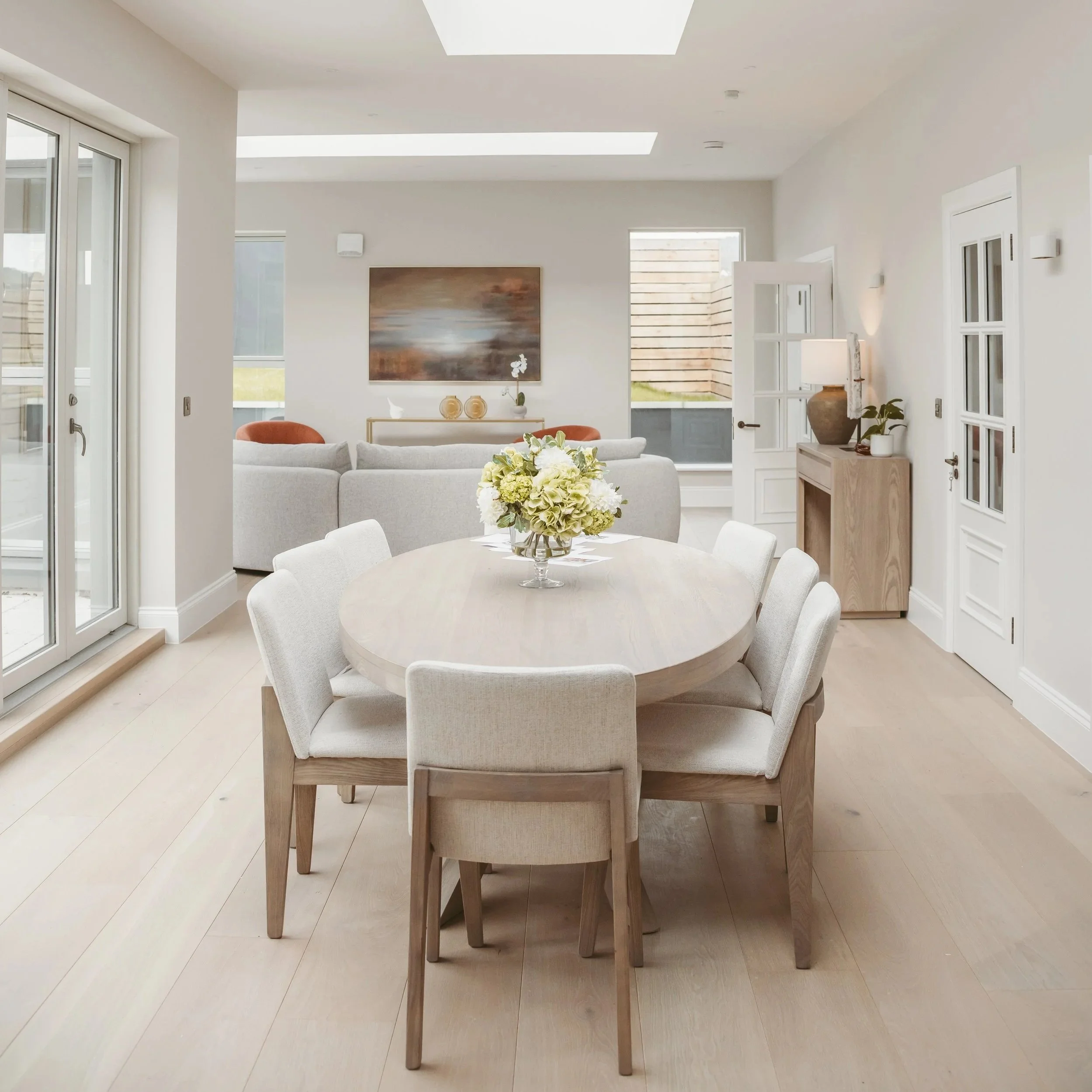 A modern, open-concept living and dining room area with white walls and large windows, featuring a round dining table with beige chairs, a gray sofa, a large abstract painting, and decorative vases and plants.