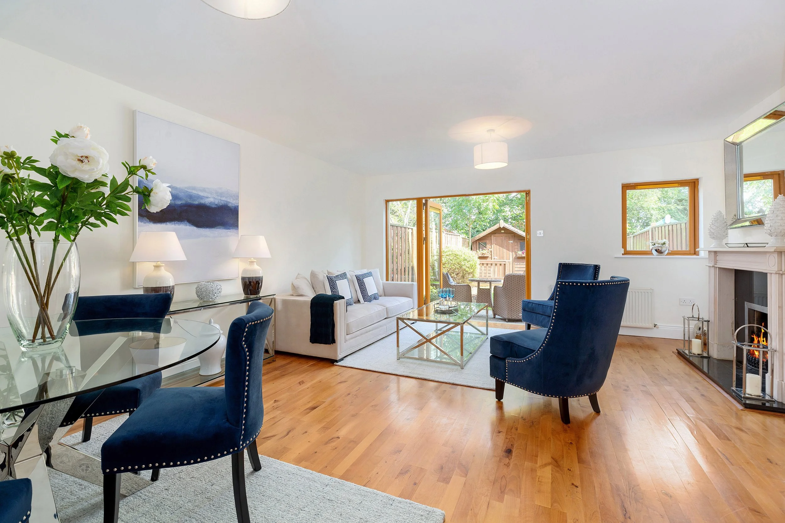 Bright living room with white walls, large windows, and hardwood floors. Contains white sofa, blue velvet armchairs, glass coffee table, and decorative lamps. Outdoor patio visible through open door.