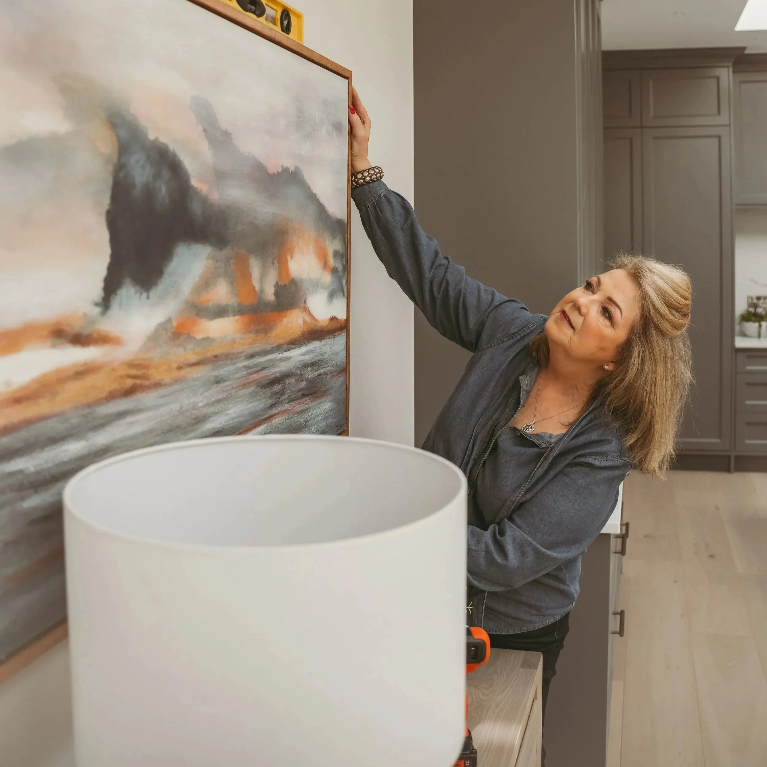 Woman with blonde hair and a black top placing artwork on a wall in a modern kitchen.