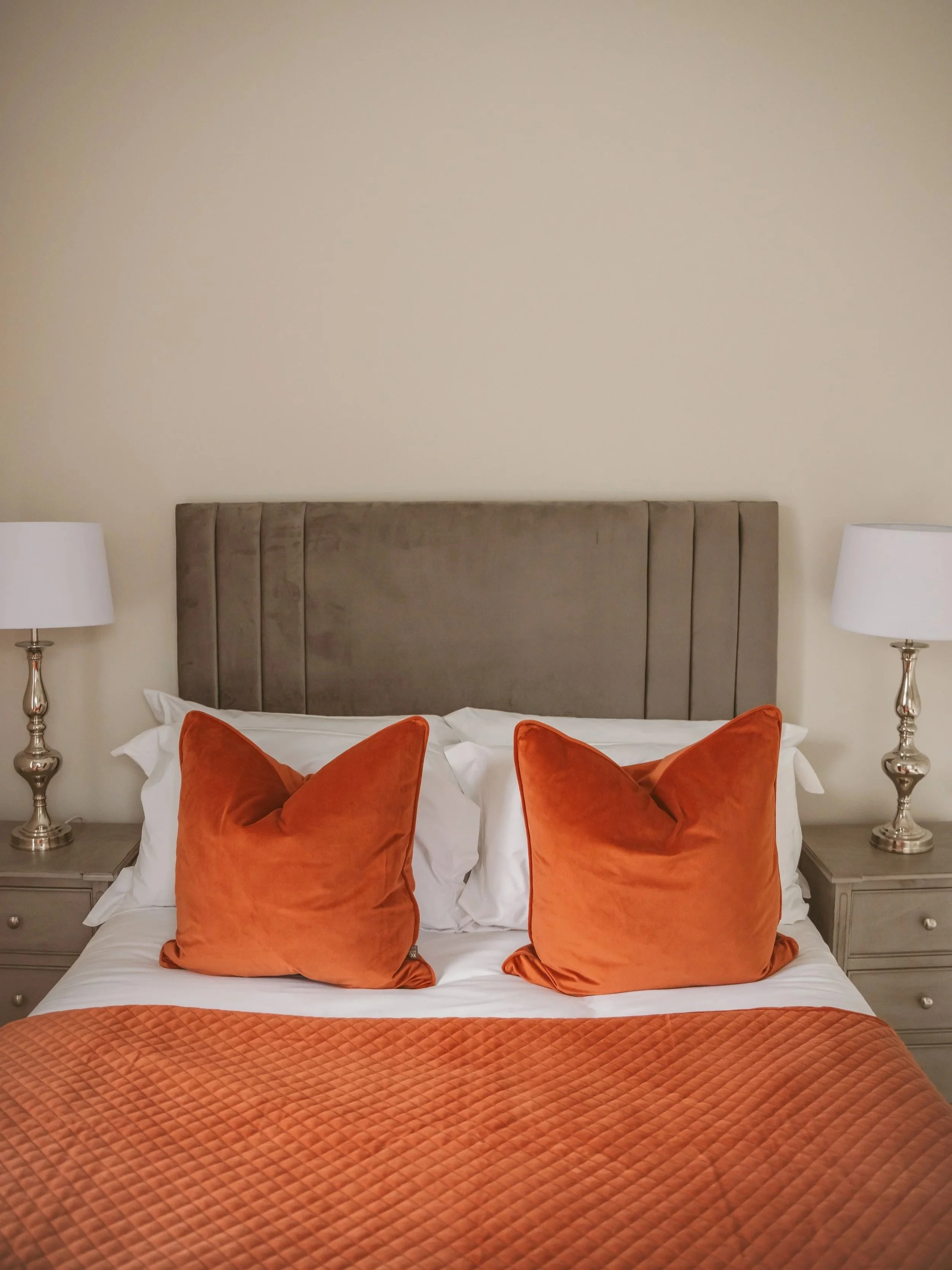 A neatly made bed with an upholstered headboard, two orange throw pillows, white sheets, orange quilted bedspread, flanked by two matching nightstands with white lamps, against a cream-colored wall.