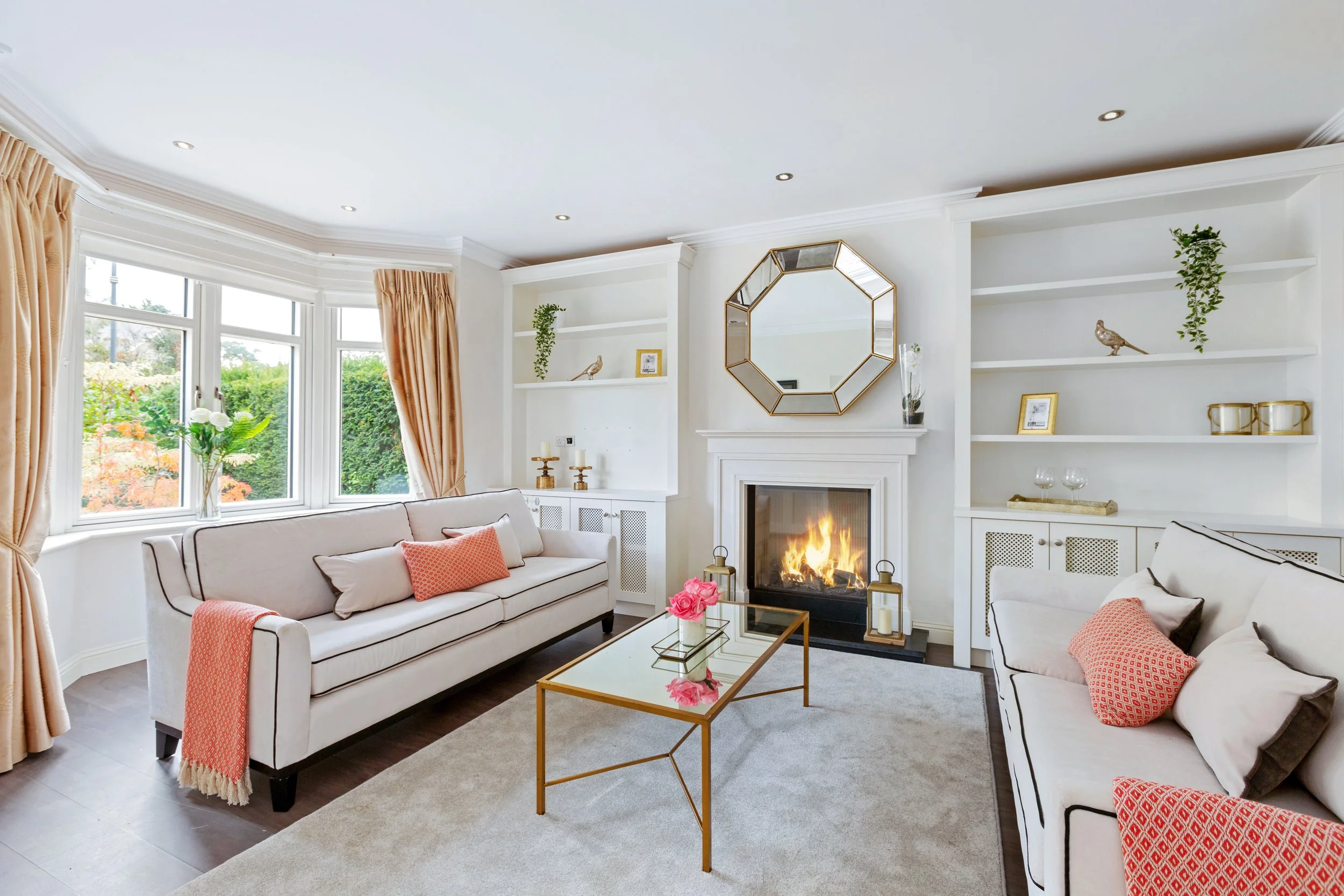 A bright living room with large bay windows, white sofas with pink and brown cushions, a white fireplace, and built-in bookshelves decorated with plants, framed photos, and decorative items. There's a gold-framed octagonal mirror on the wall and a glass coffee table with pink flowers in the center.
