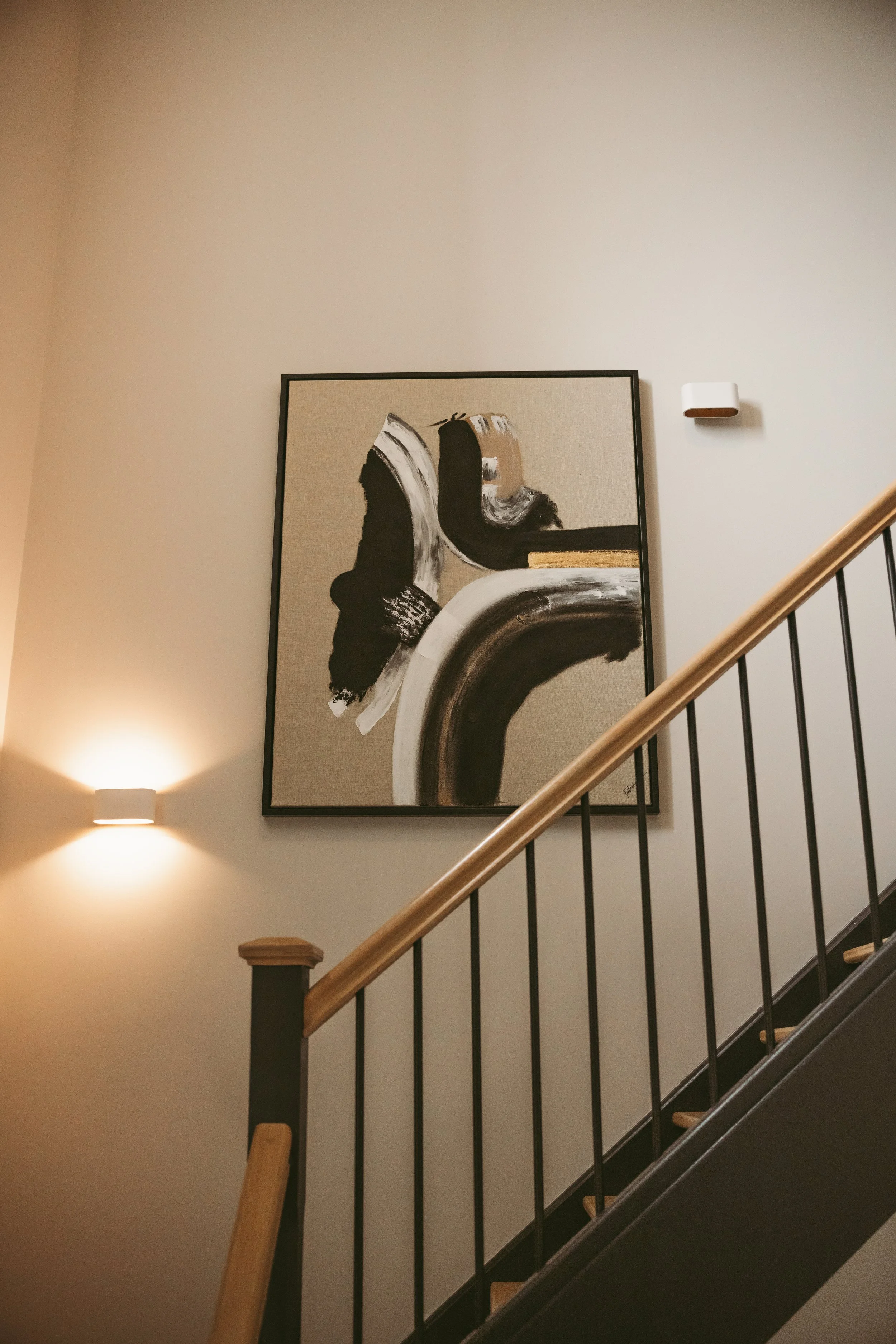Interior staircase with a painting on the wall, a wall sconce light, and a handrail.
