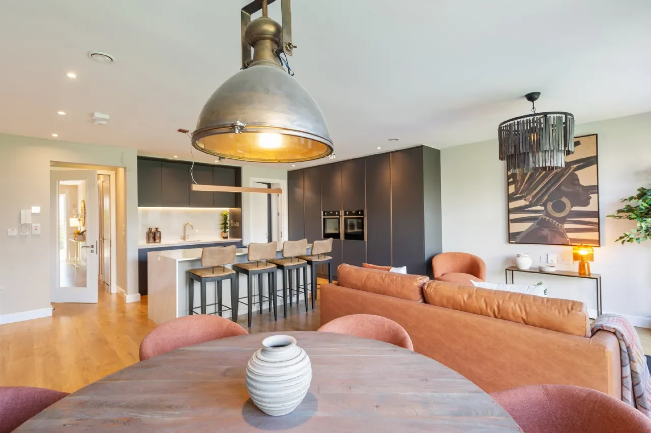 Open-concept living room and kitchen with modern decor, featuring a round wooden dining table with a white vase, tan and pink chairs, a leather sofa, and various artworks and lighting fixtures.