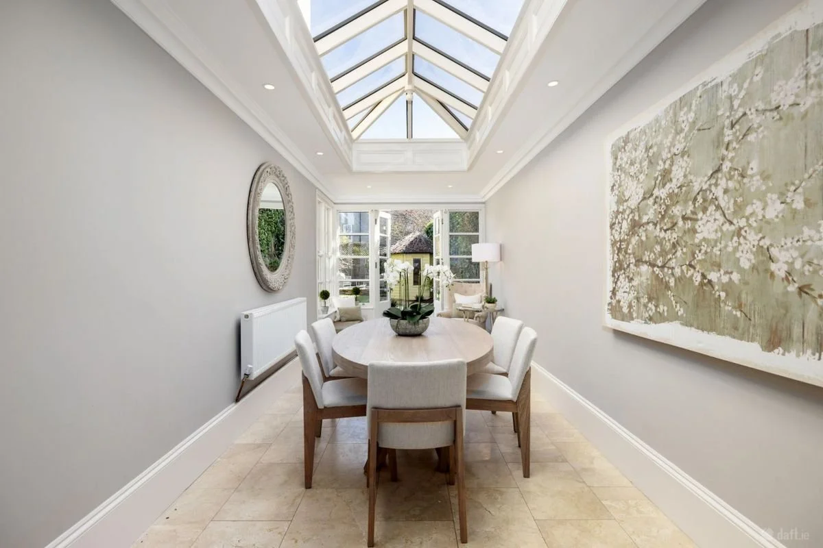 Bright dining room with a skylight roof, white walls, and a wooden table with six white upholstered chairs, large window at the back, and decorative art on the walls.