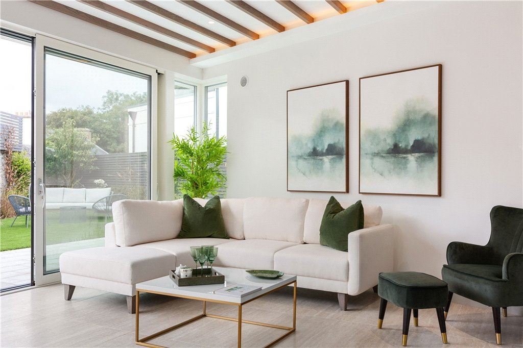 A bright living room with a white sectional sofa, green and black pillows, a marble-top coffee table with glassware, a black armchair with small matching ottoman, a large potted plant, and framed watercolor landscape art on the wall. Sliding glass door opens to a backyard with outdoor seating and greenery.