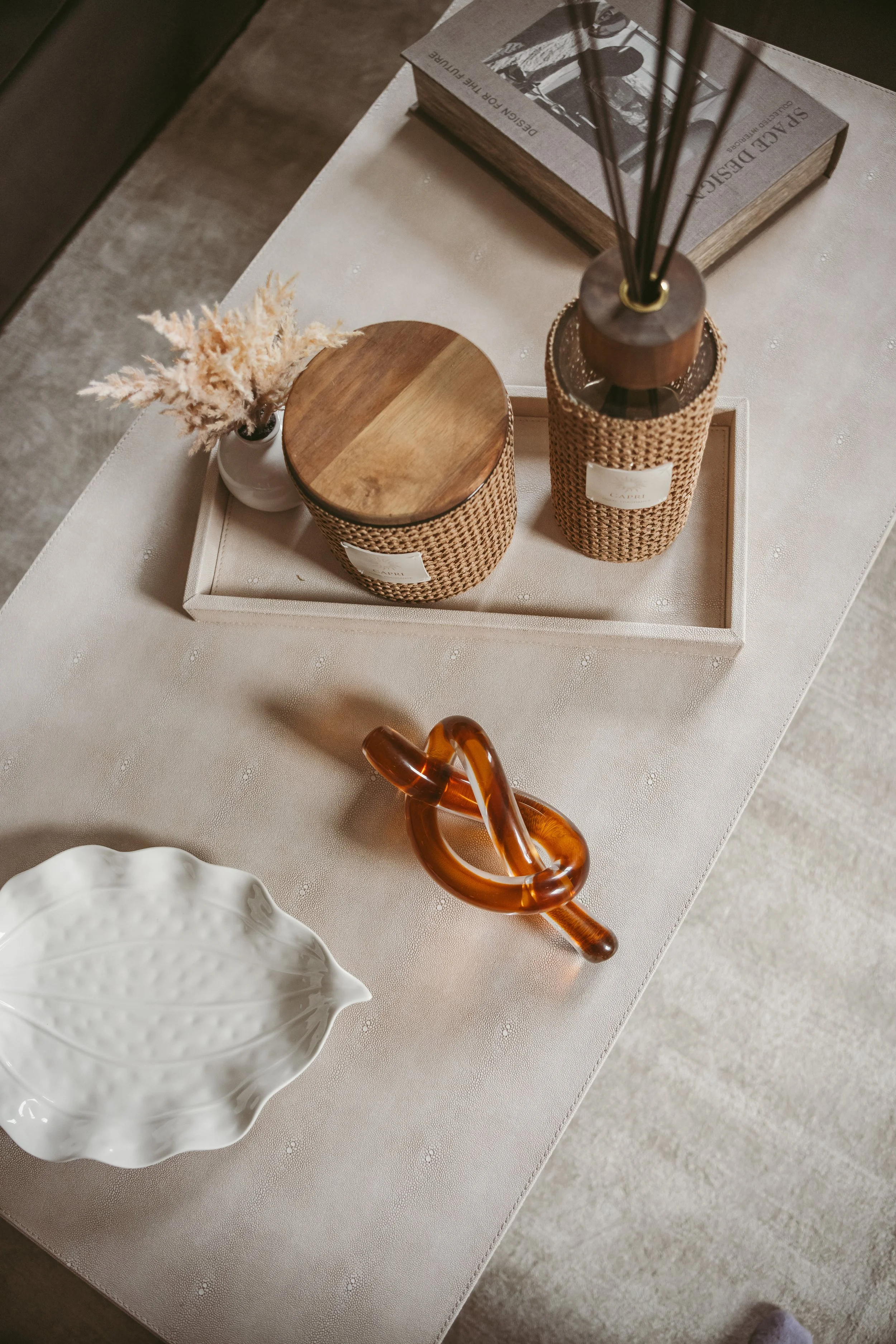 Top-down view of a decorated corner table with a book, a wicker-style perfume bottle, reed diffuser, white ceramic vase with dried flowers, and a tortoise shell shaped glass sculpture on a white textured table.