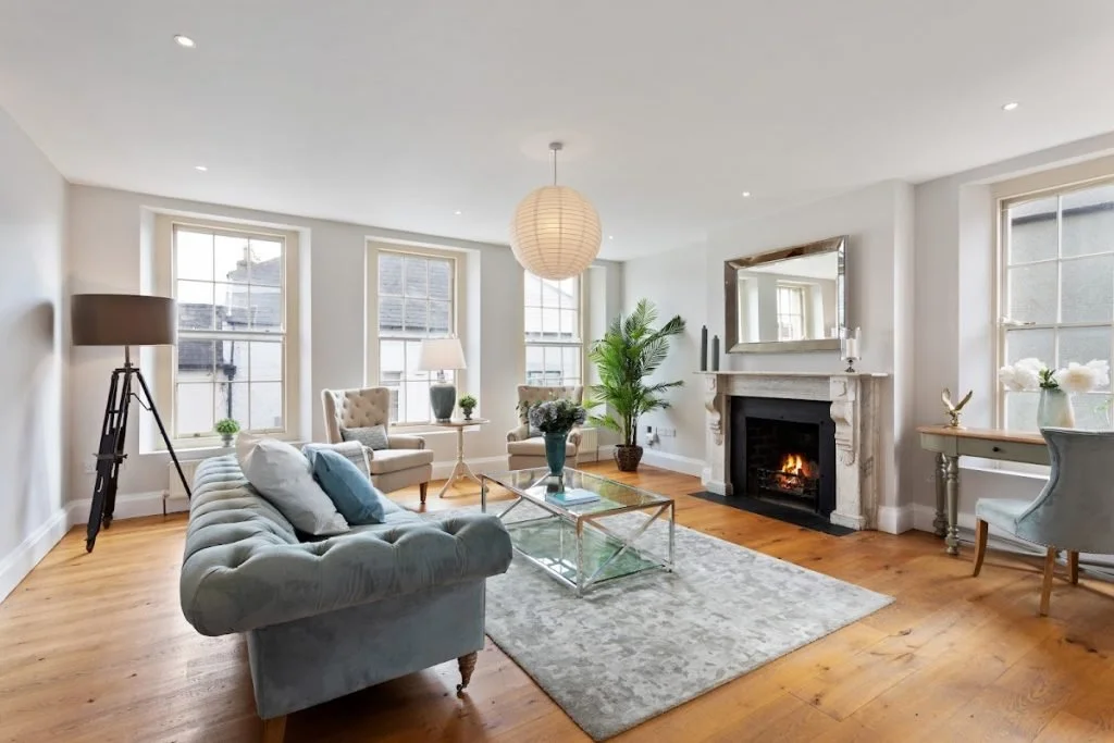 Bright living room with three large windows allowing natural light, white walls, hardwood floors, a gray tufted sofa with throw pillows, a glass coffee table, an area rug, a white fireplace with a mirror above, a large potted plant, a side table with a lamp, and various chairs and decorative items.