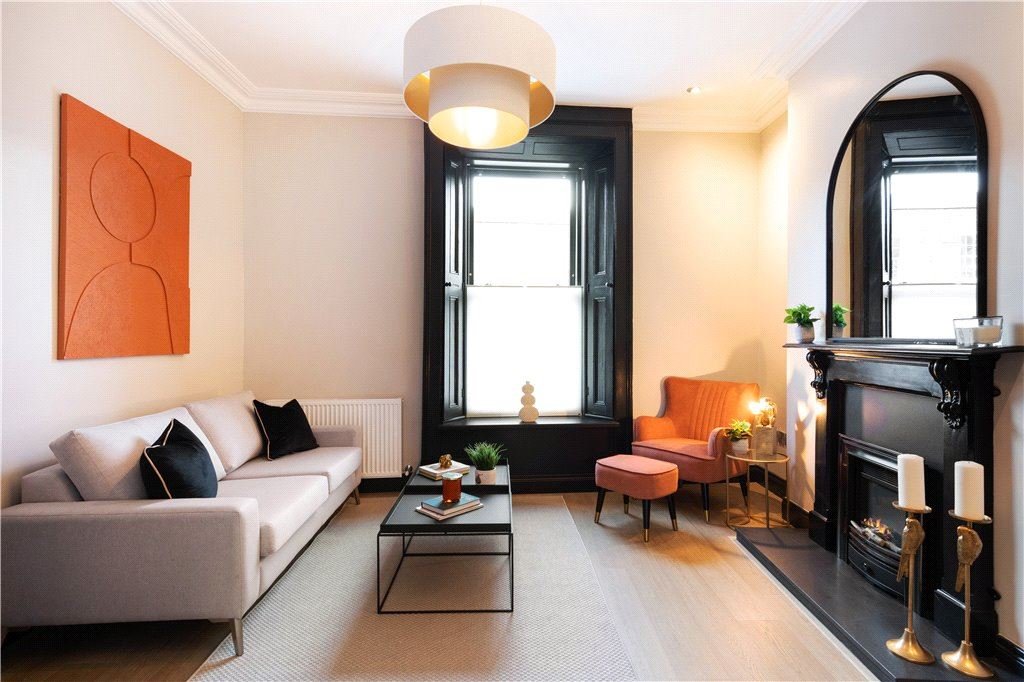 Living room with a white sofa, black pillows, a coffee table, a large window with black trim, a pink armchair with a matching ottoman, a gold side table, a black fireplace with candles, and a wall mirror.
