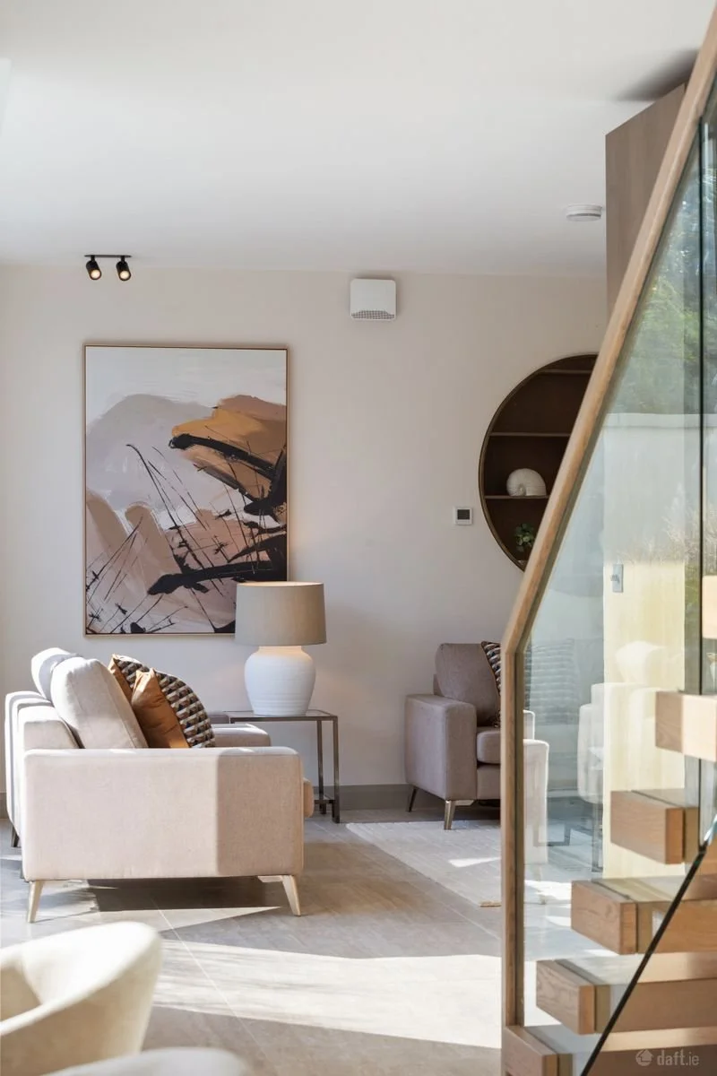 A modern living room with light-colored sofa, armchair, abstract art on the wall, table lamp, and decorative shelf, featuring neutral tones and natural light.
