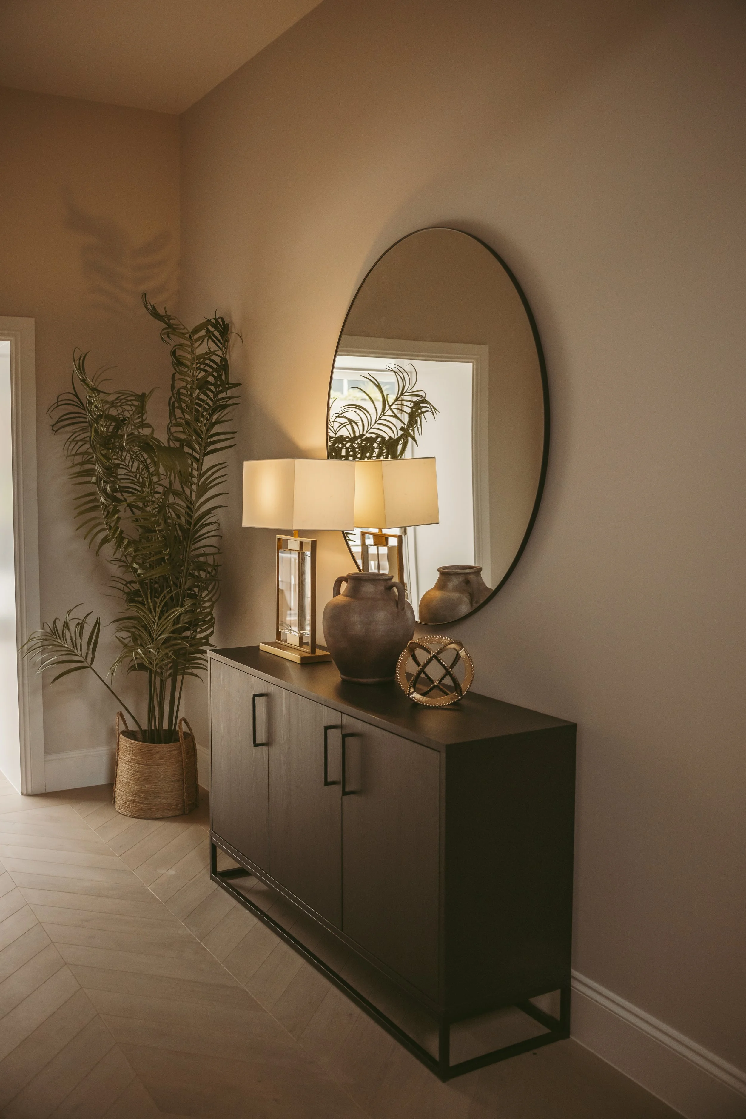 A black sideboard with decorative items, including two lamps, ceramic vases, and a gold wire sculpture, positioned against a light-colored wall. A large, oval mirror hangs above the sideboard, reflecting part of a room with a window and ceiling light. To the left, there is a tall potted plant in a woven basket.