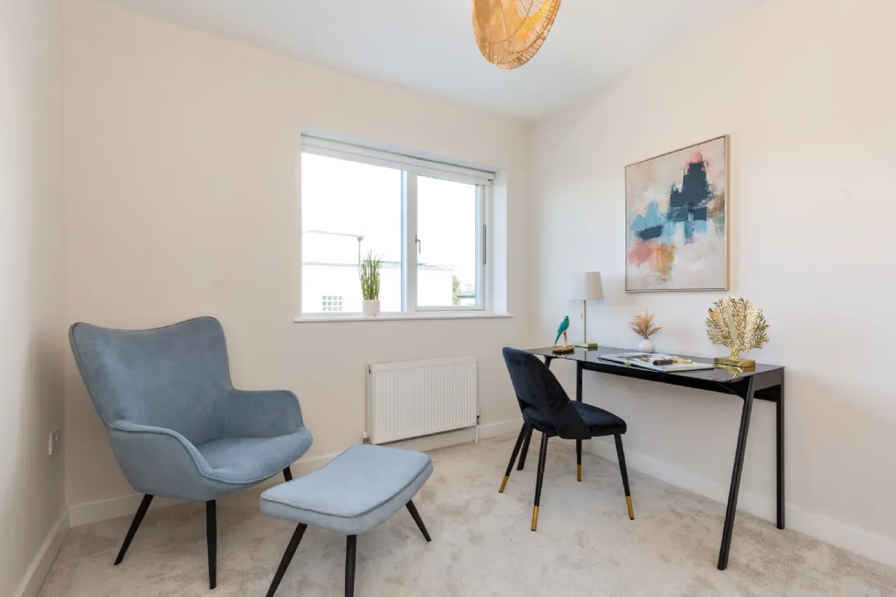 A minimalist home office with a window, a blue armchair with a matching ottoman, a black desk with a black chair, a table lamp, and decorative objects on the desk, including a small plant and art.