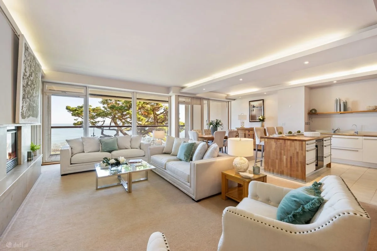 Bright living room with white sofas, large window view of trees, and an open kitchen with island.