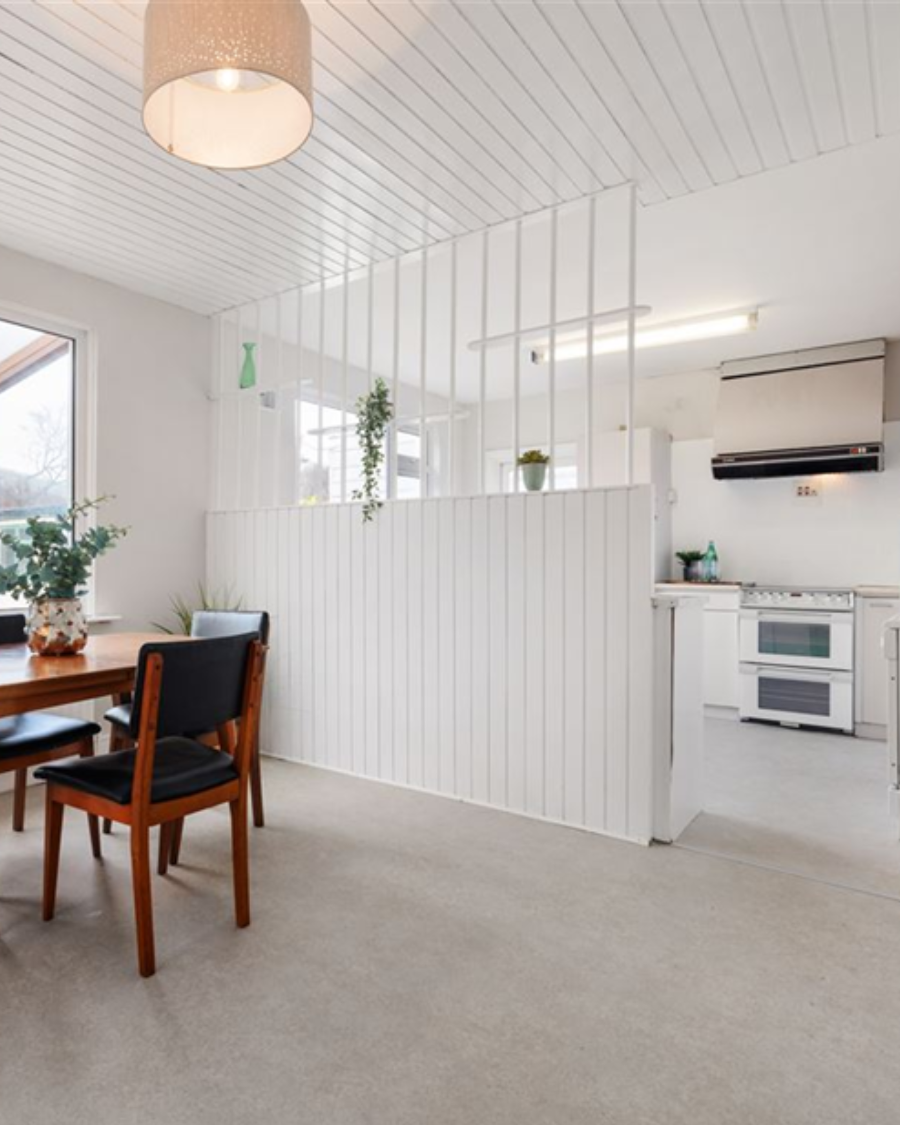 A dining area with a wooden table and black chairs, decorated with potted plants, next to windows that let in natural light. In the background, there is a white kitchen with an oven, stove, and wall-mounted microwave, separated by a white slatted partial wall. The ceiling is white with wood paneling and features a beige hanging light fixture.