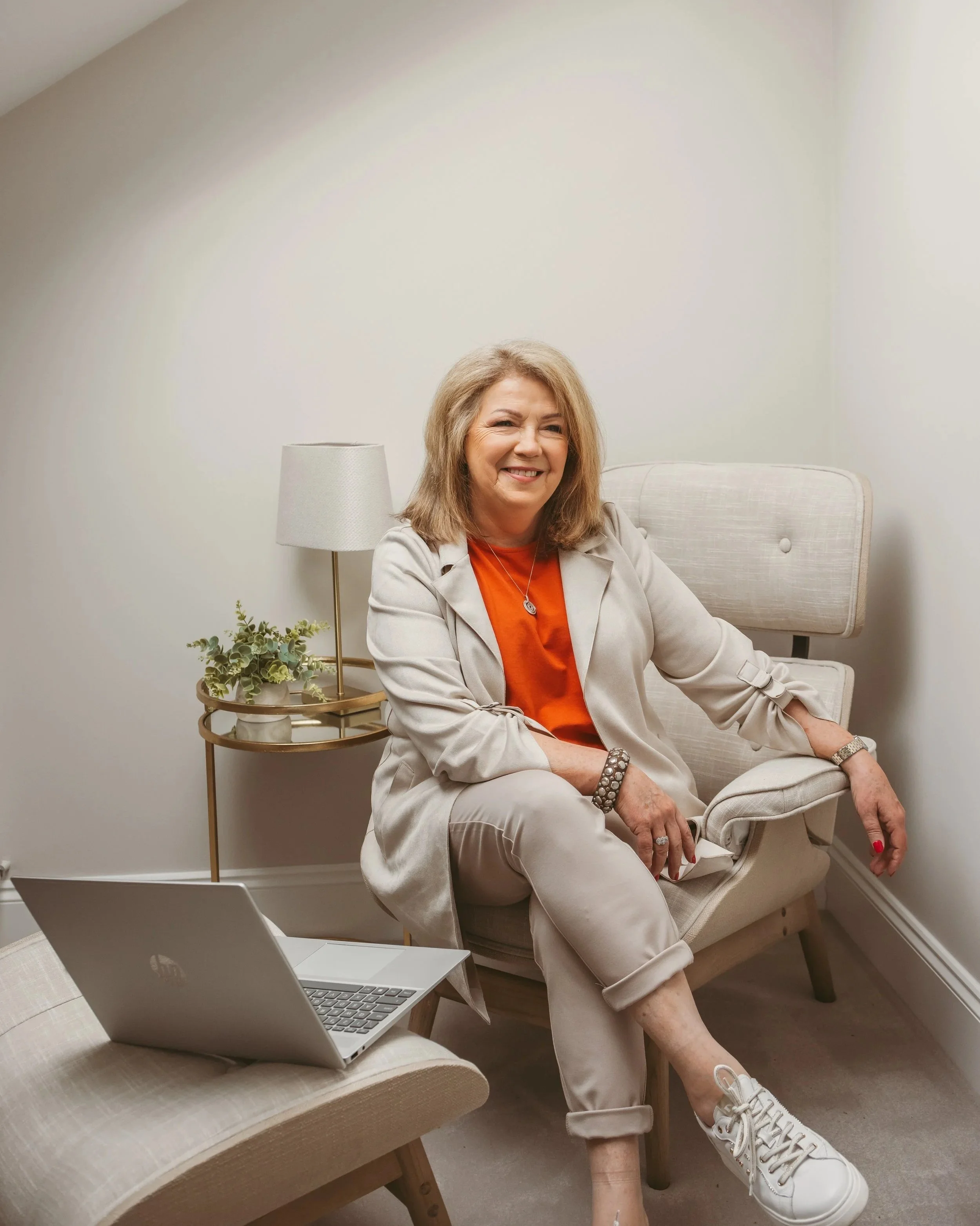 Woman sitting in a beige armchair, smiling and winking, with a laptop on a beige table in front of her, in a cozy room with white walls, a beige lamp, and greenery on the side.