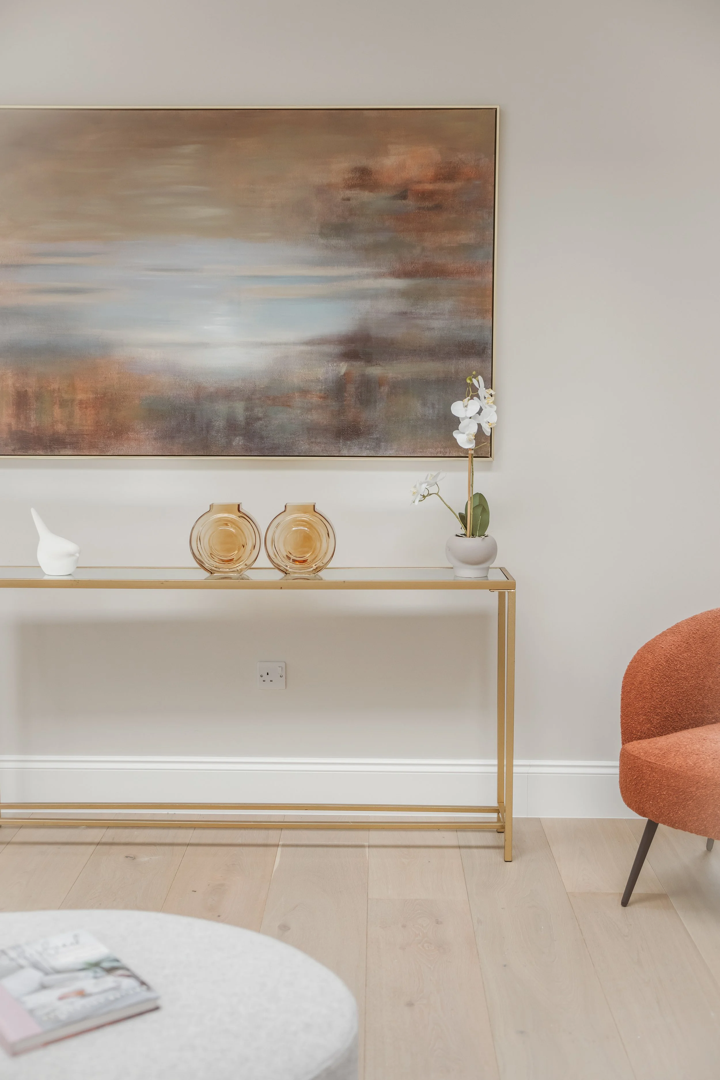 Minimalist interior decor with a large abstract painting, a white orchid in a vase, decorative glass vases on a glass console table, and a reddish-orange upholstered chair.