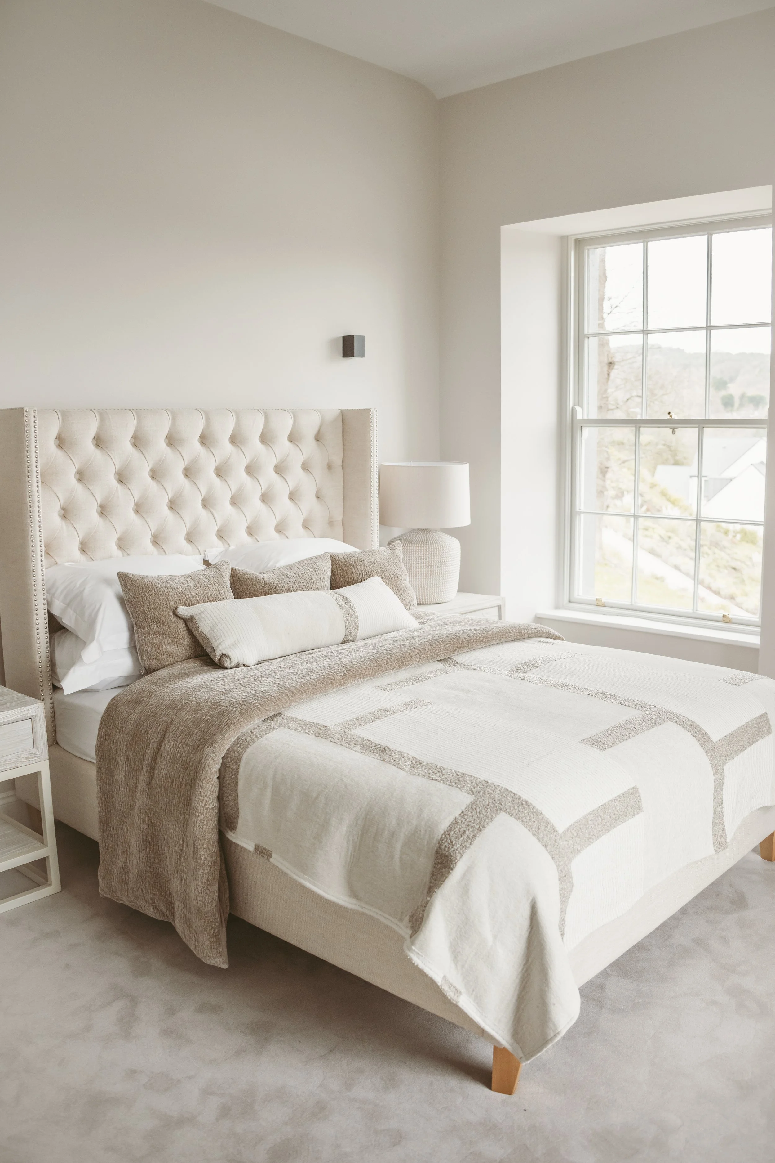 A neatly made bed with a beige and white patterned bedspread, beige pillows, and a beige throw blanket. The bed has a cream-colored, tufted, upholstered headboard. Next to the bed is a white nightstand with a woven ceramic lamp. A large window on the right allows natural light into the room, showing a view of houses and trees outside.