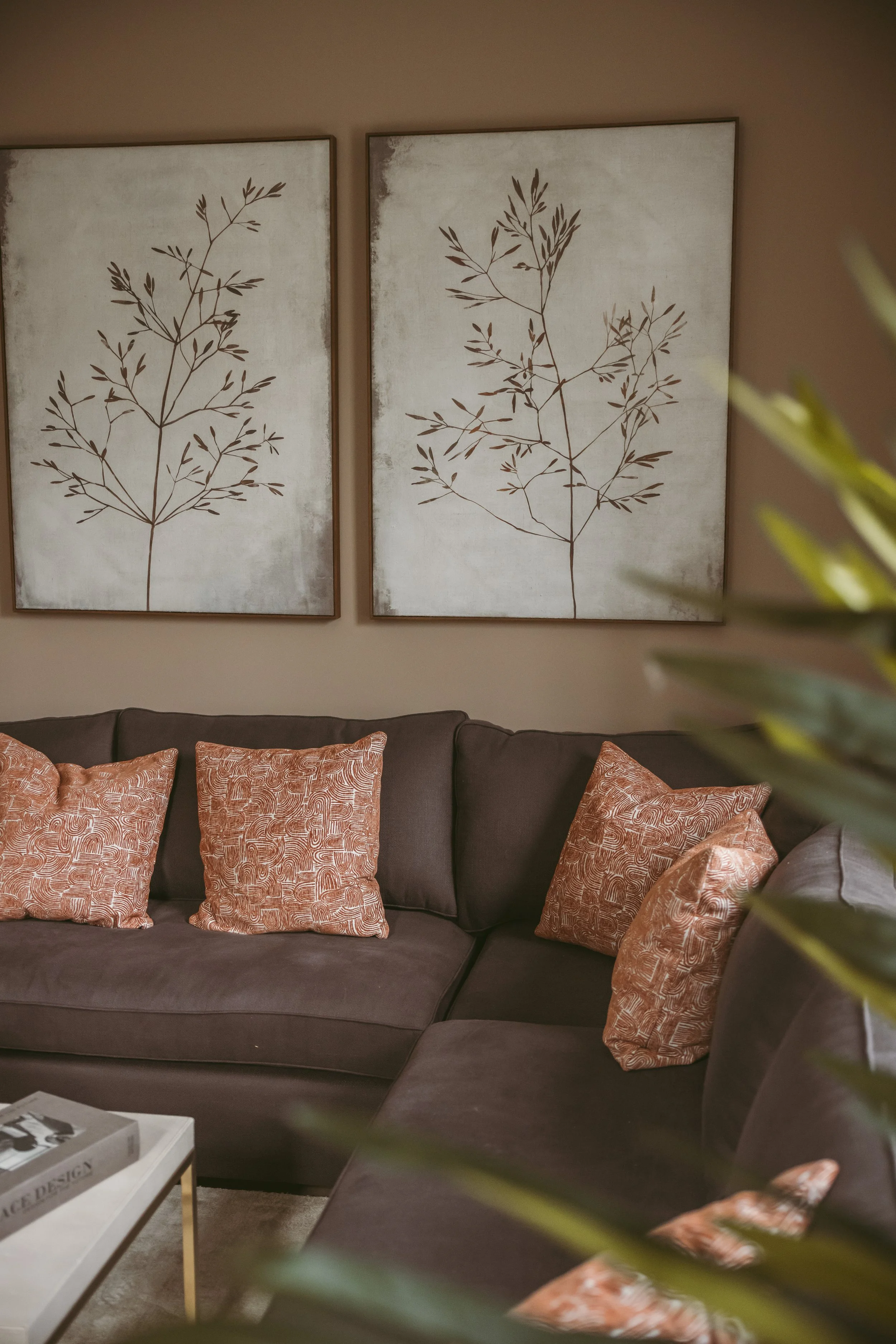Living room with dark grey sofa and multiple orange patterned pillows, white coffee table with a book, and two large wall art pieces featuring brown botanical sketches on a beige background.