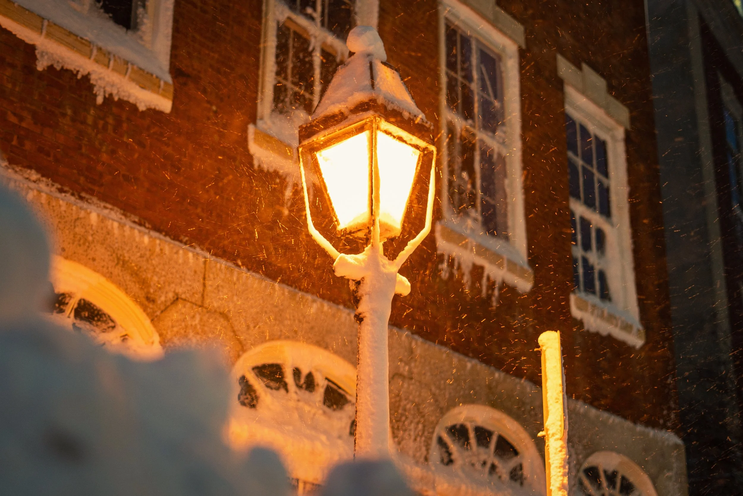 A snow-covered street lamp illuminating a snowy street at night with a brick building in the background.