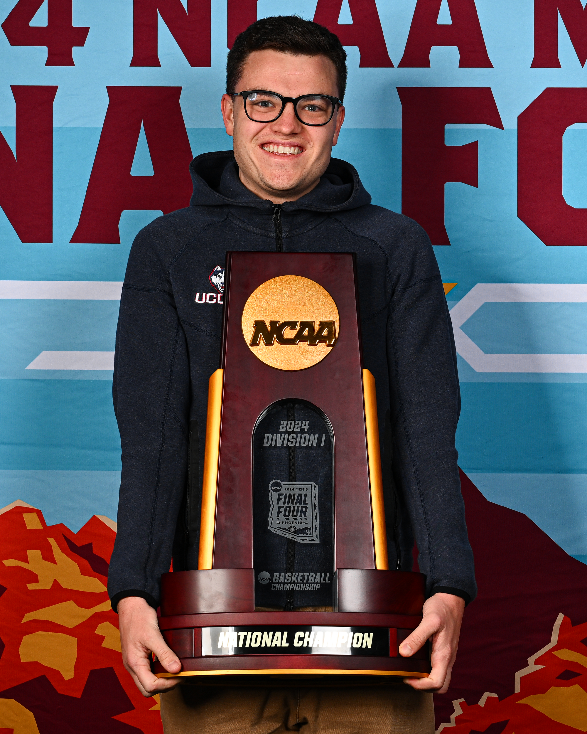 Young man holding trophy with NCAA logo, smiling, wearing glasses and a UConn hoodie, standing in front of a backdrop with NCAA branding and mountains.