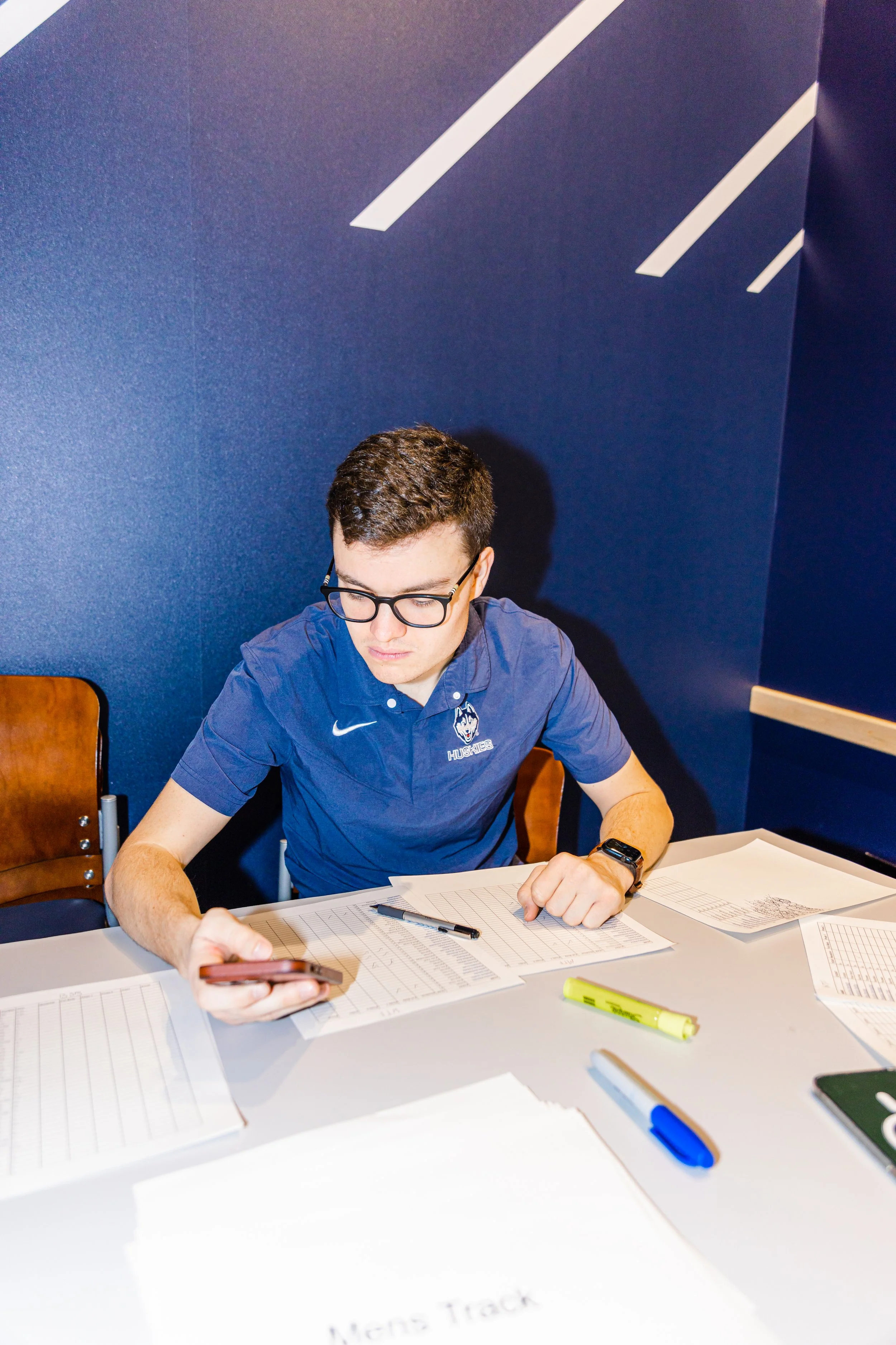 A young man with glasses, wearing a navy blue polo shirt, is sitting at a table covered with papers, with a pen, highlighters, and a smartphone in his hand, focused on his work against a dark blue wall.