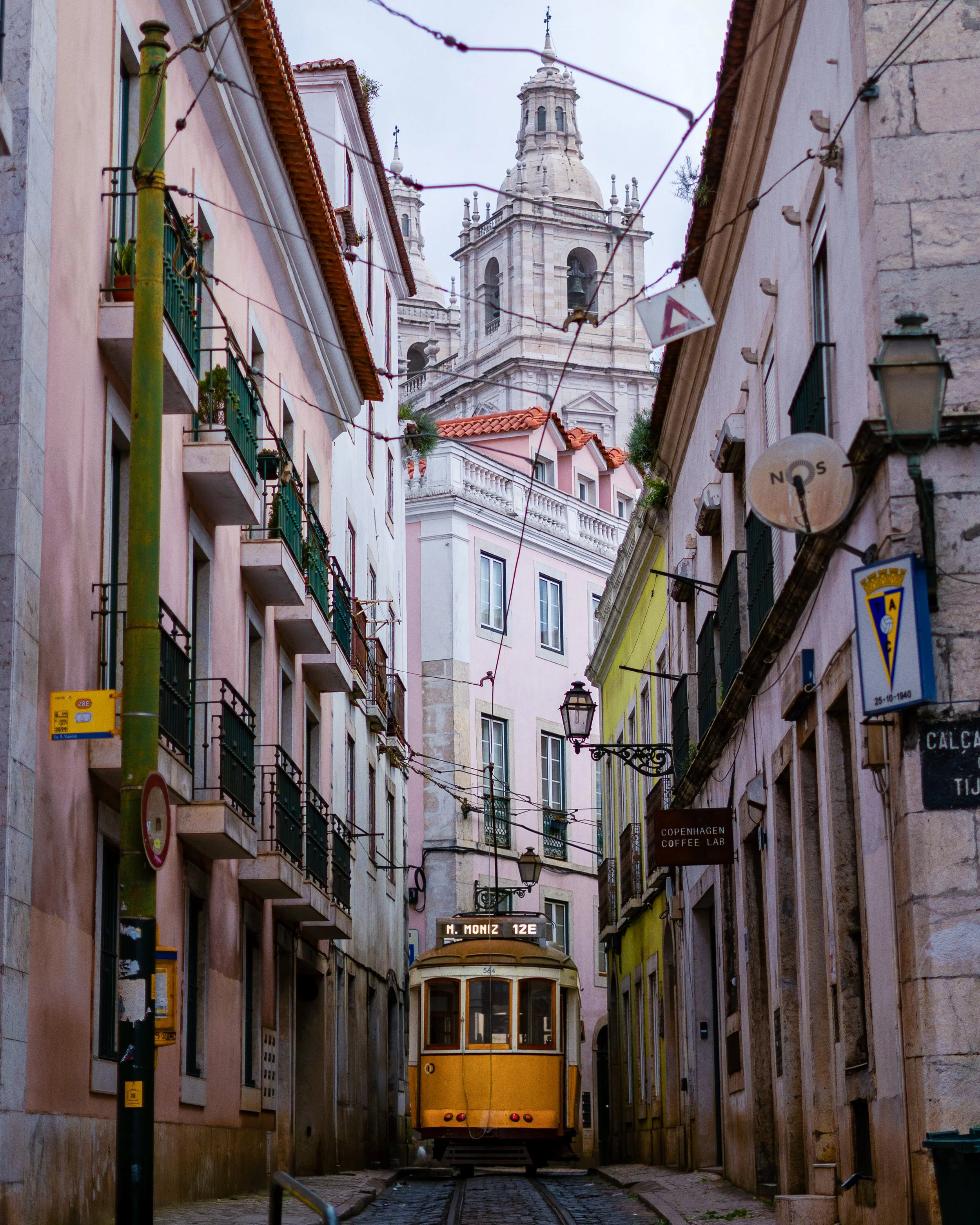 A yellow tram on a narrow city street surrounded by pastel-colored buildings with balconies and a church tower in the background.
