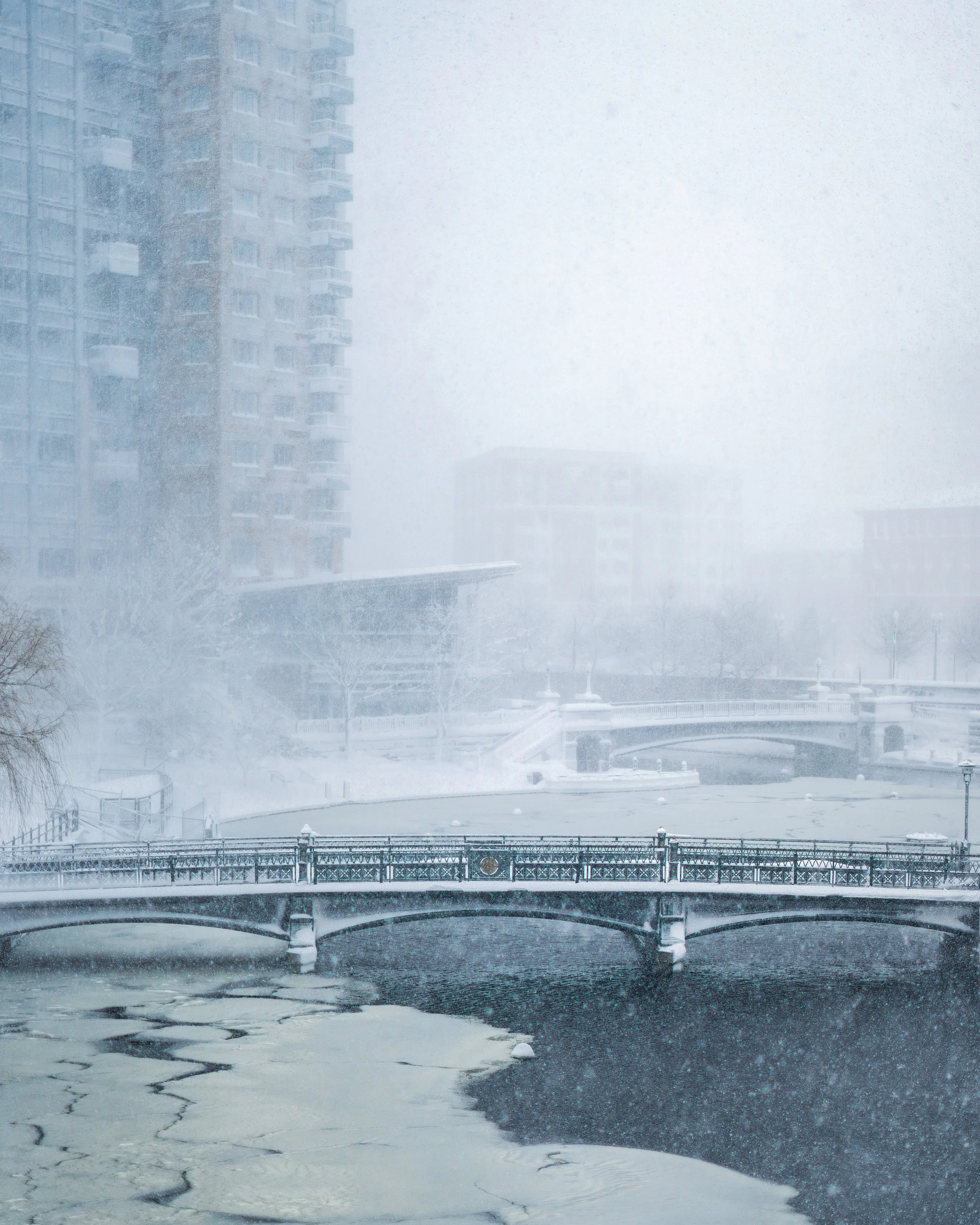 A winter city scene with high-rise buildings, a bridge over a partially frozen river, and snow falling heavily, creating a foggy atmosphere.