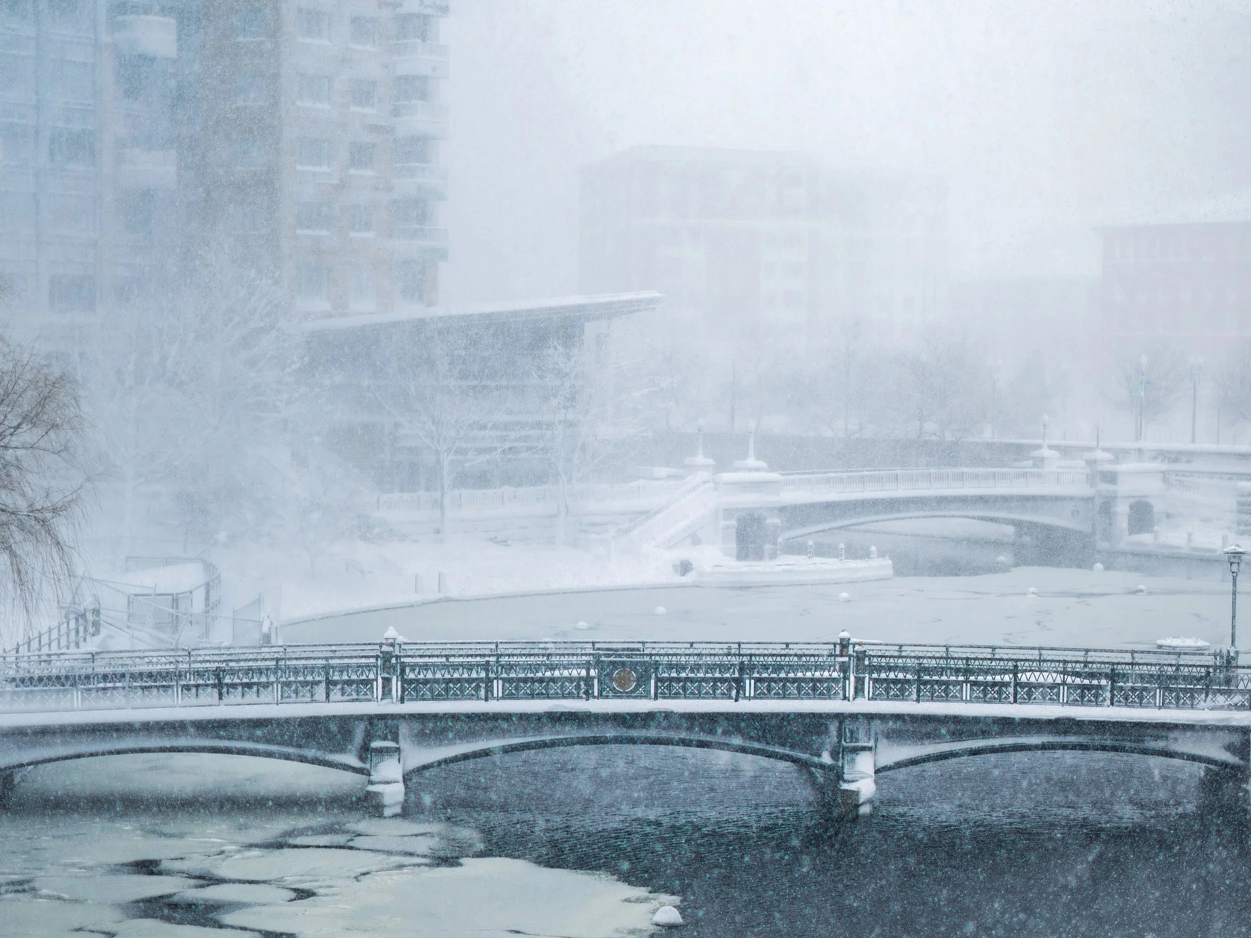 Snow-covered bridge over a partially frozen river with snow-covered trees and buildings in the background during a snowstorm.