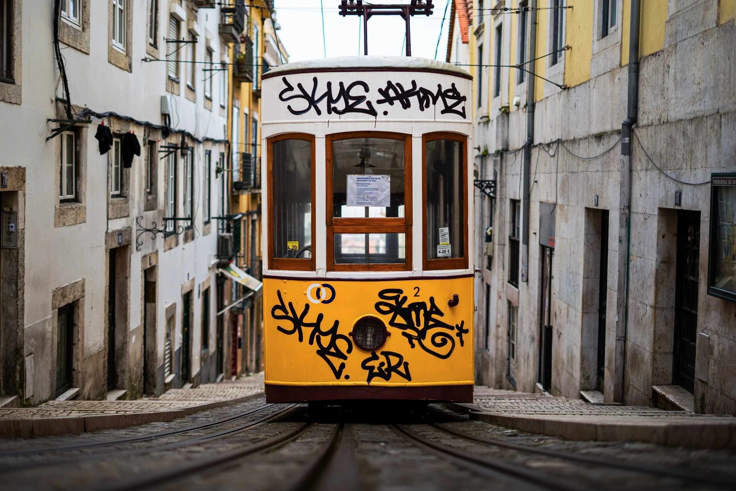 A yellow vintage tram with graffiti on its front, traveling down a narrow street with tall buildings on either side.