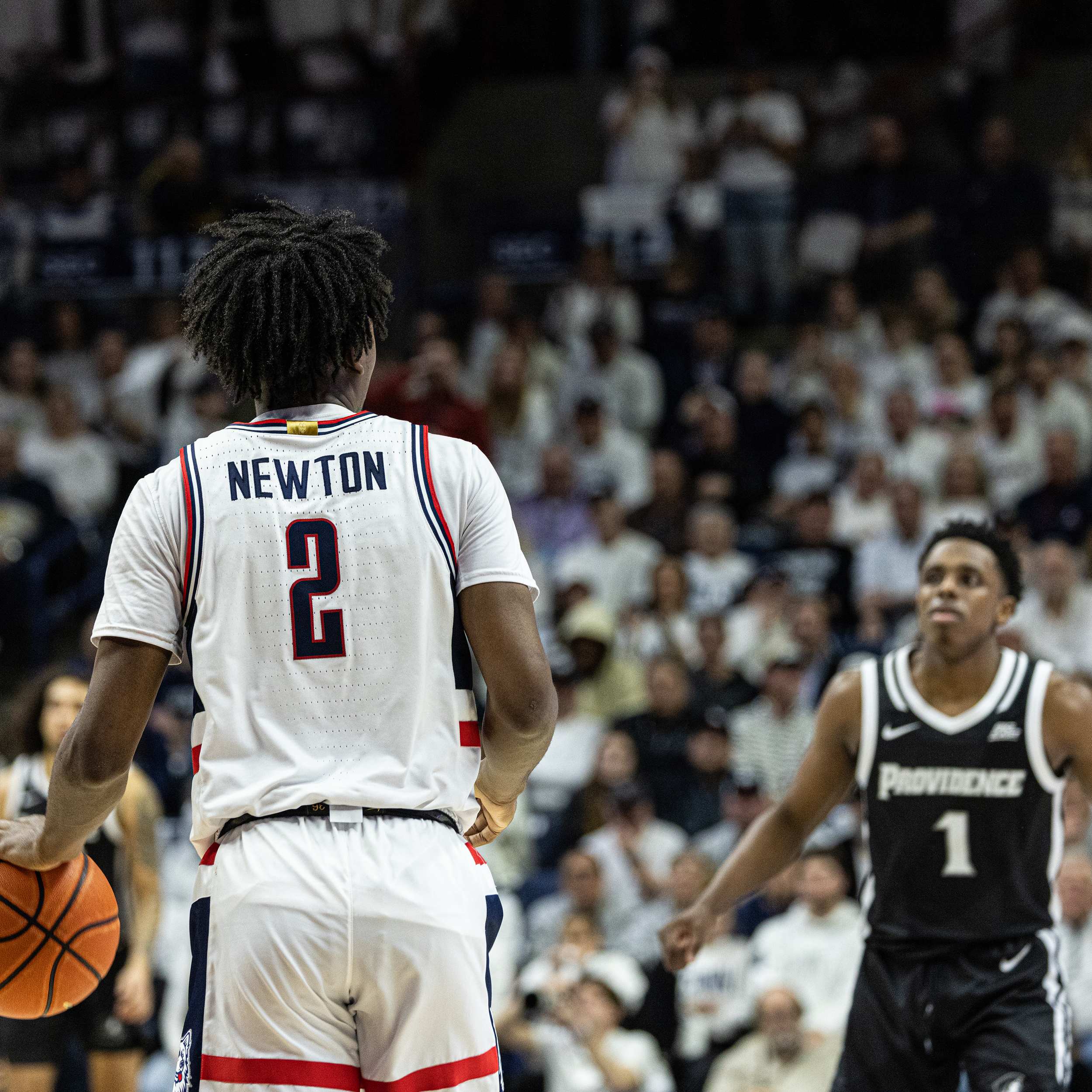 Back of a basketball player in a white jersey with red and blue accents, number 2, with the name Newton, holding a basketball, facing another player in a black jersey, number 1, with 'Providence' written on it, on a basketball court with crowd in the background.