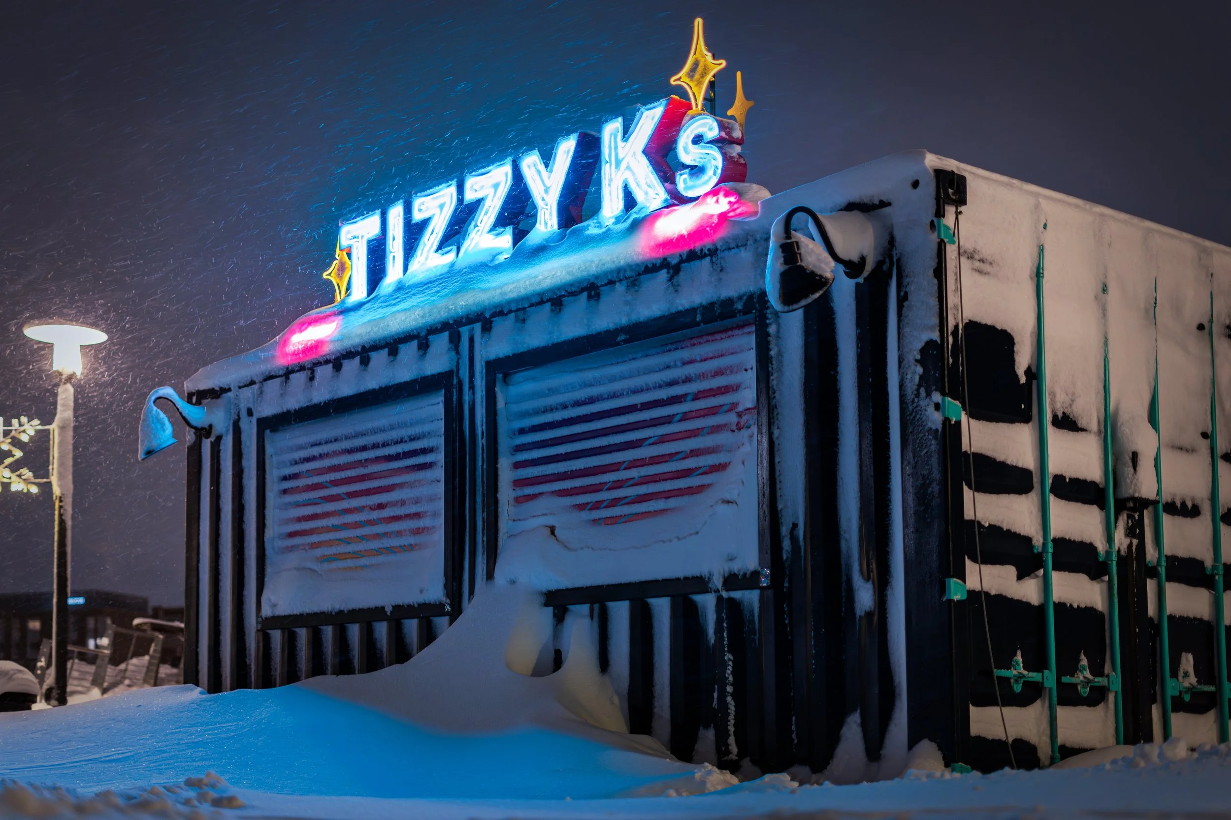 Snow-covered building with illuminated sign reading 'TIZZYS' on top, snow on the ground, snow falling, and a nearby streetlamp in the background.
