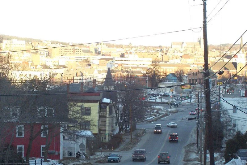 View of a hilly urban neighborhood with houses, a commercial area, and multiple cars on a winding street, seen through utility wires.