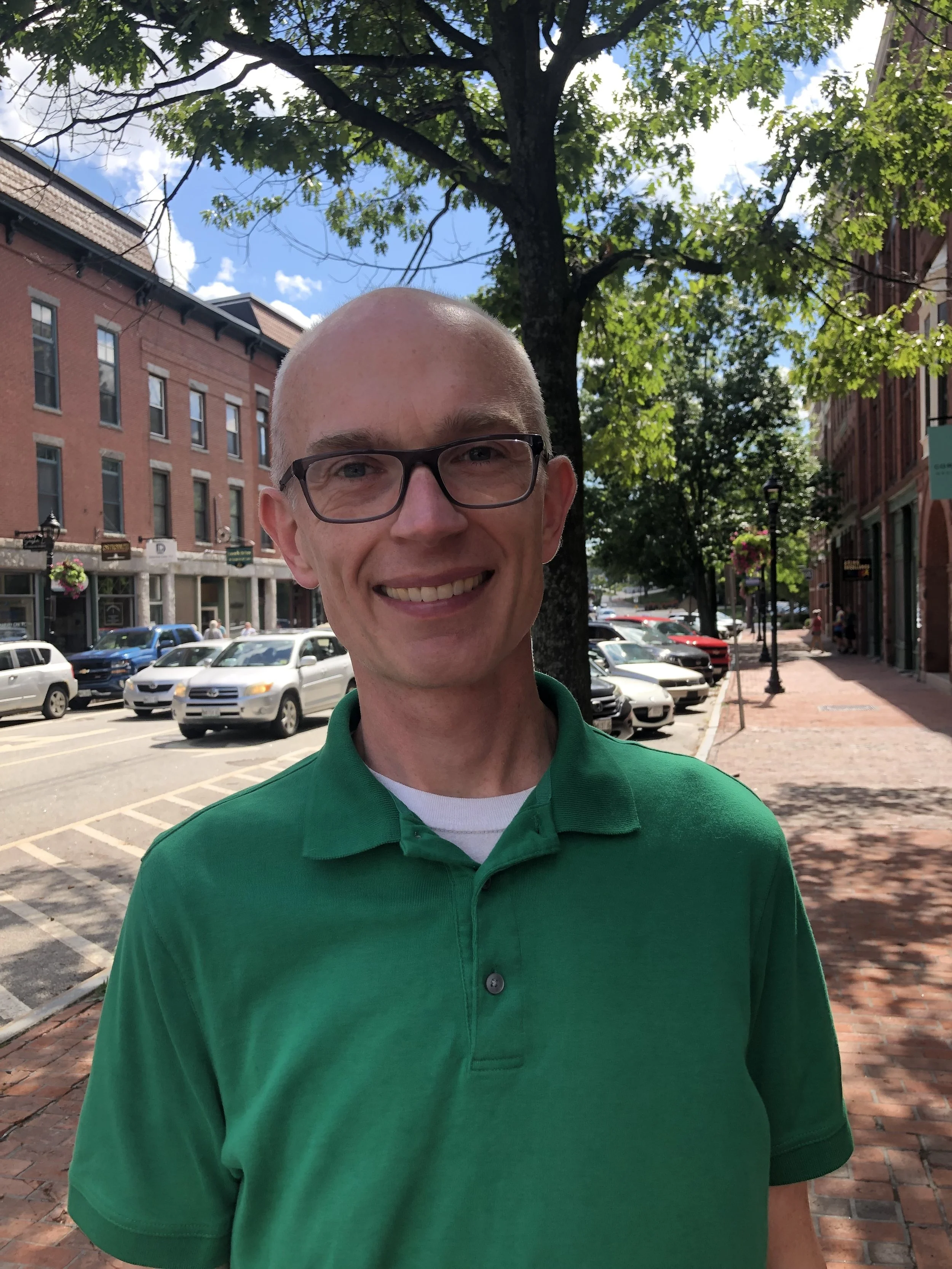 Dana Staples Auburn Maine House of Representatives district 88 A smiling man with glasses wearing a green polo shirt stands on a brick sidewalk in a downtown area, with parked cars, historic brick buildings, trees.