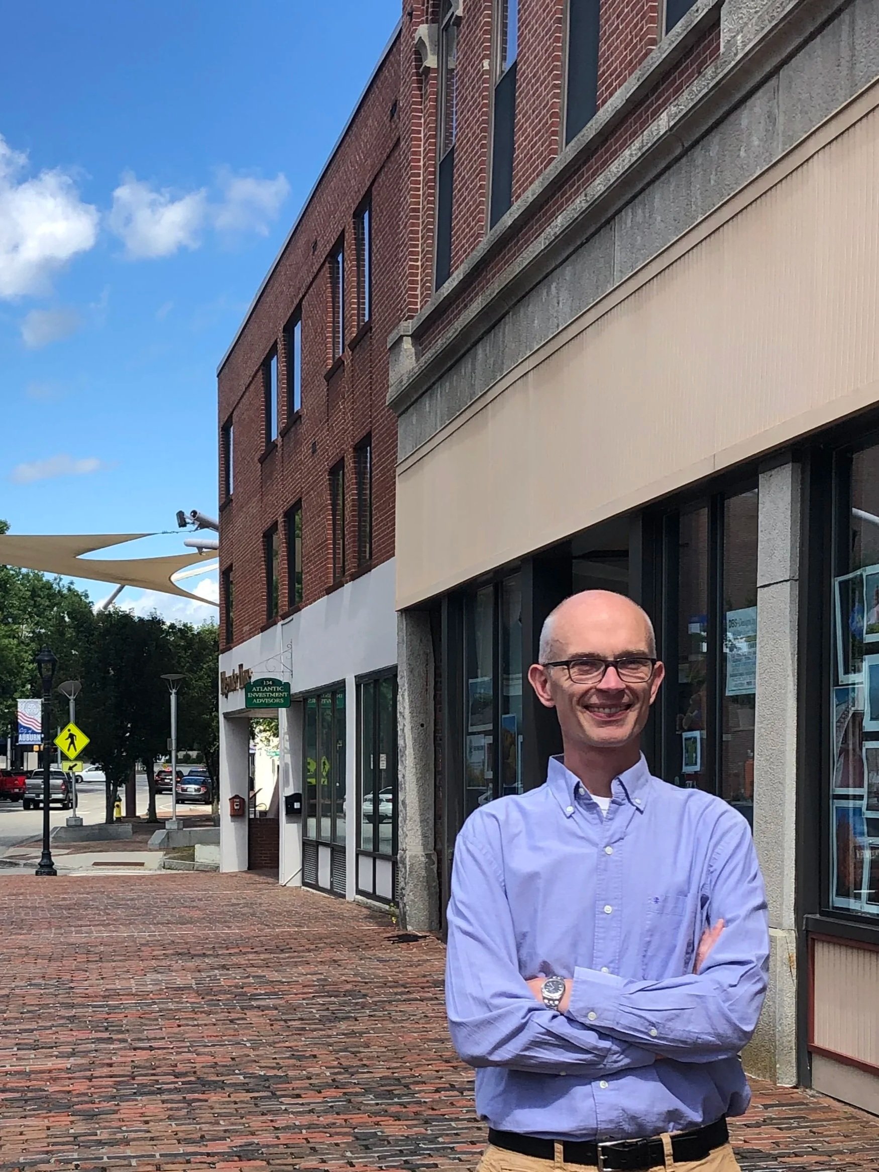 Dana Staples Auburn Maine House of Representatives district 88 A man with glasses and a blue button-up shirt standing outdoors on a brick sidewalk in front of a brick building with large glass windows and a beige awning.