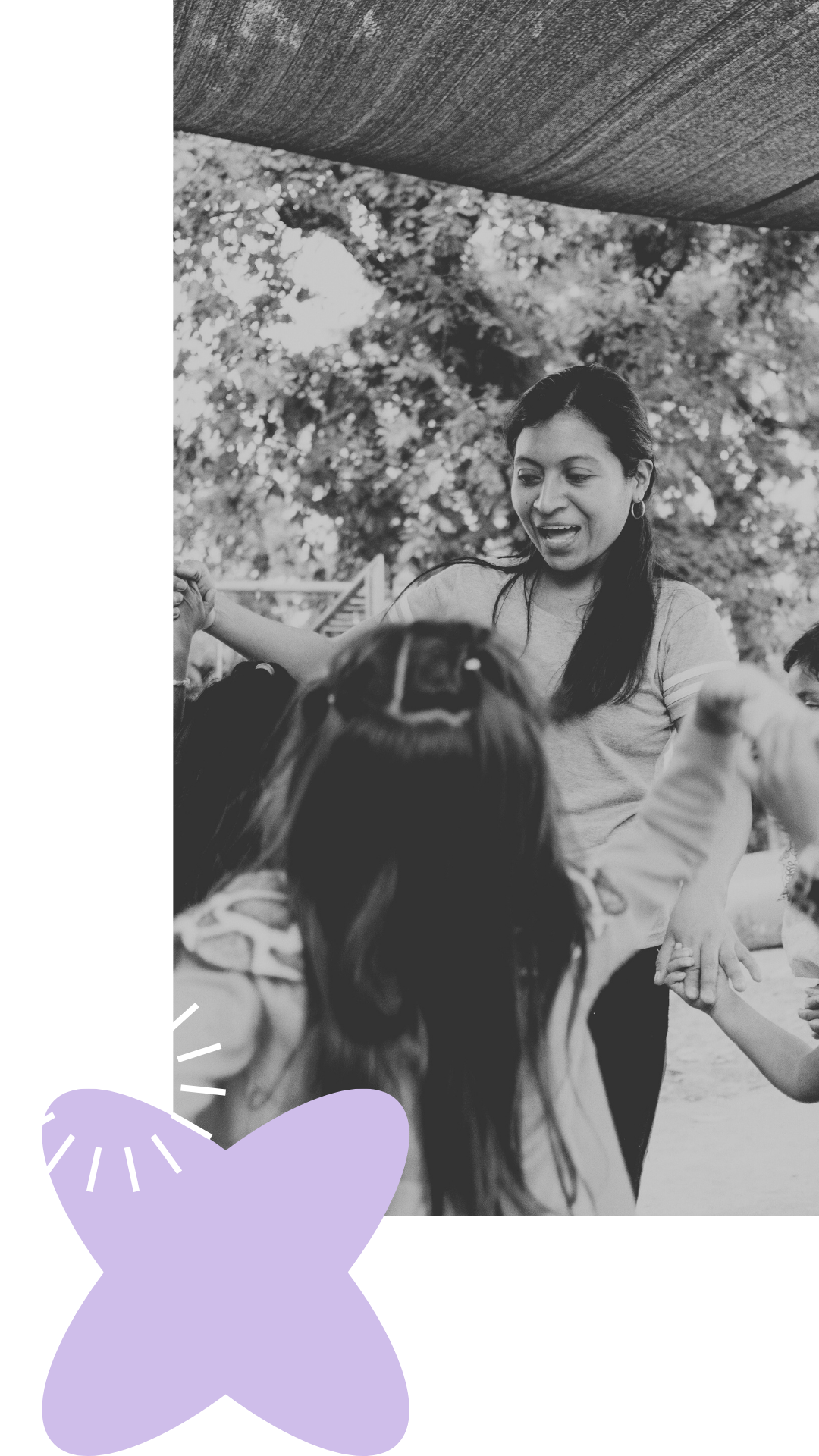 A woman with long dark hair wearing a light-colored t-shirt smiling and interacting with children outdoors under a canopy, with trees and sunlight in the background, in black and white.
